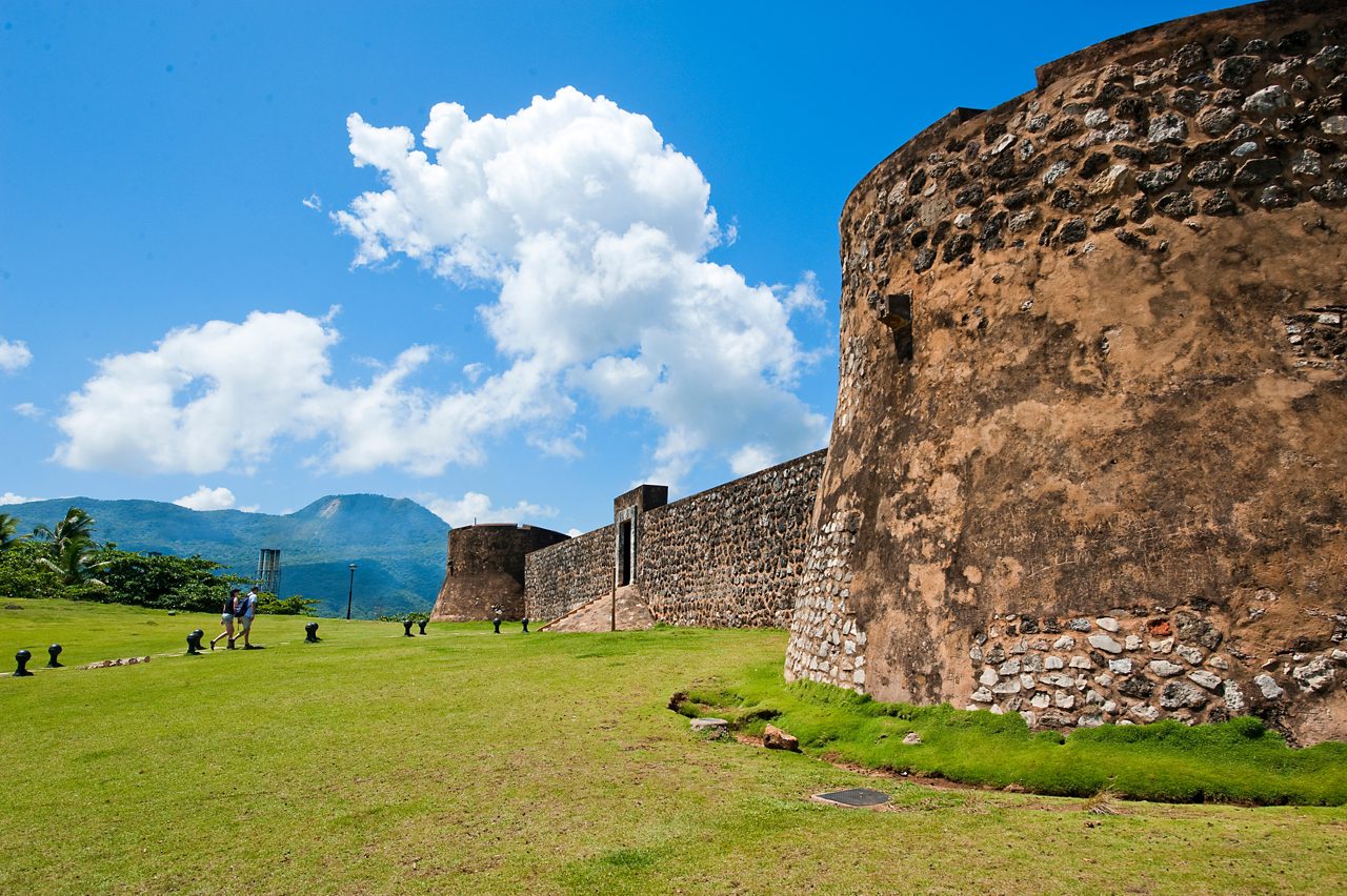 A view of Fortaleza San Felipe in Puerto Plata, Dominican Republic, featuring weathered stone fortifications surrounded by green lawns and set against a scenic mountainous backdrop with clear blue skies.