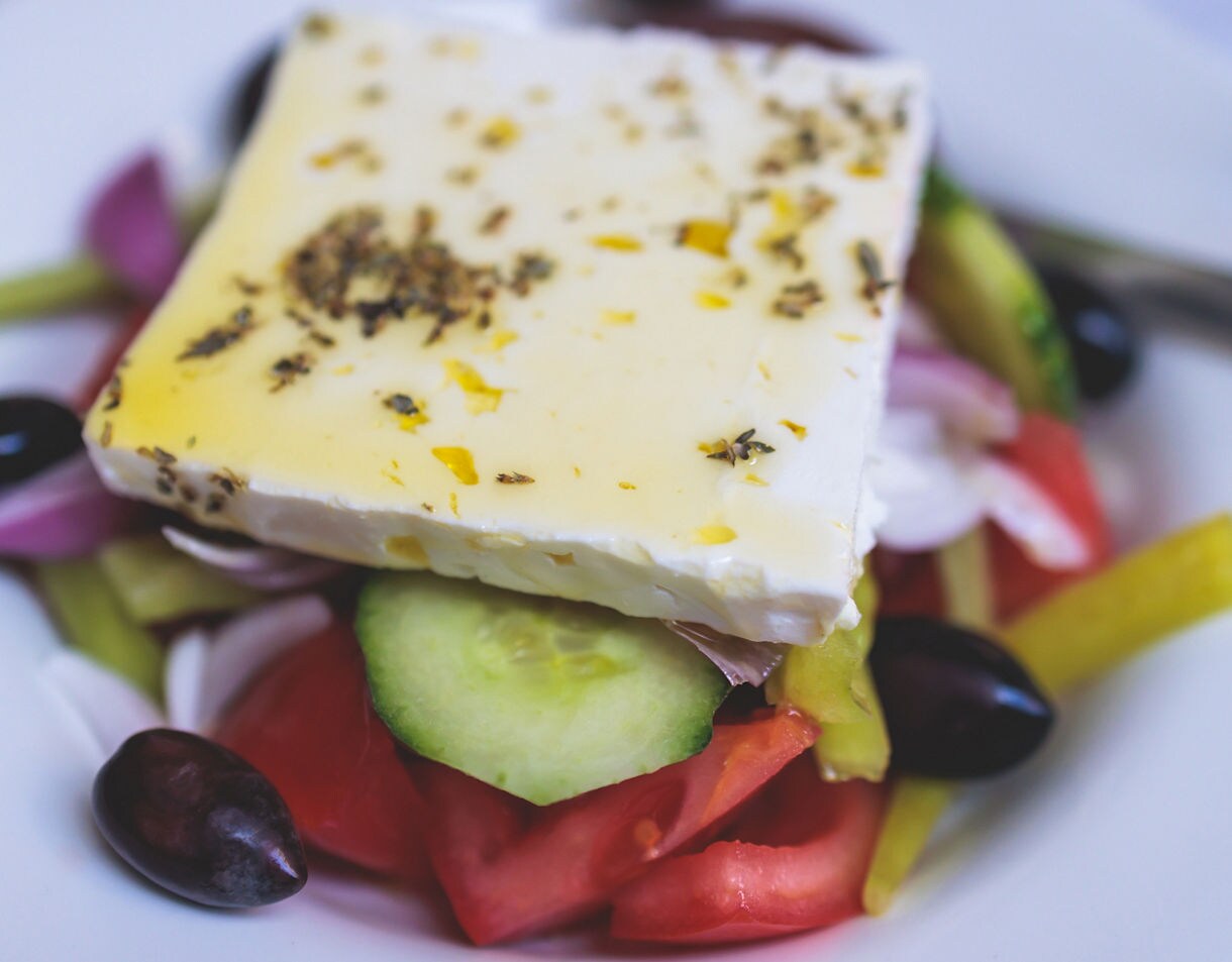 Close-up of a Greek salad topped with feta cheese, olive oil, oregano, tomatoes, cucumbers, onions and Kalamata olives on a white plate.