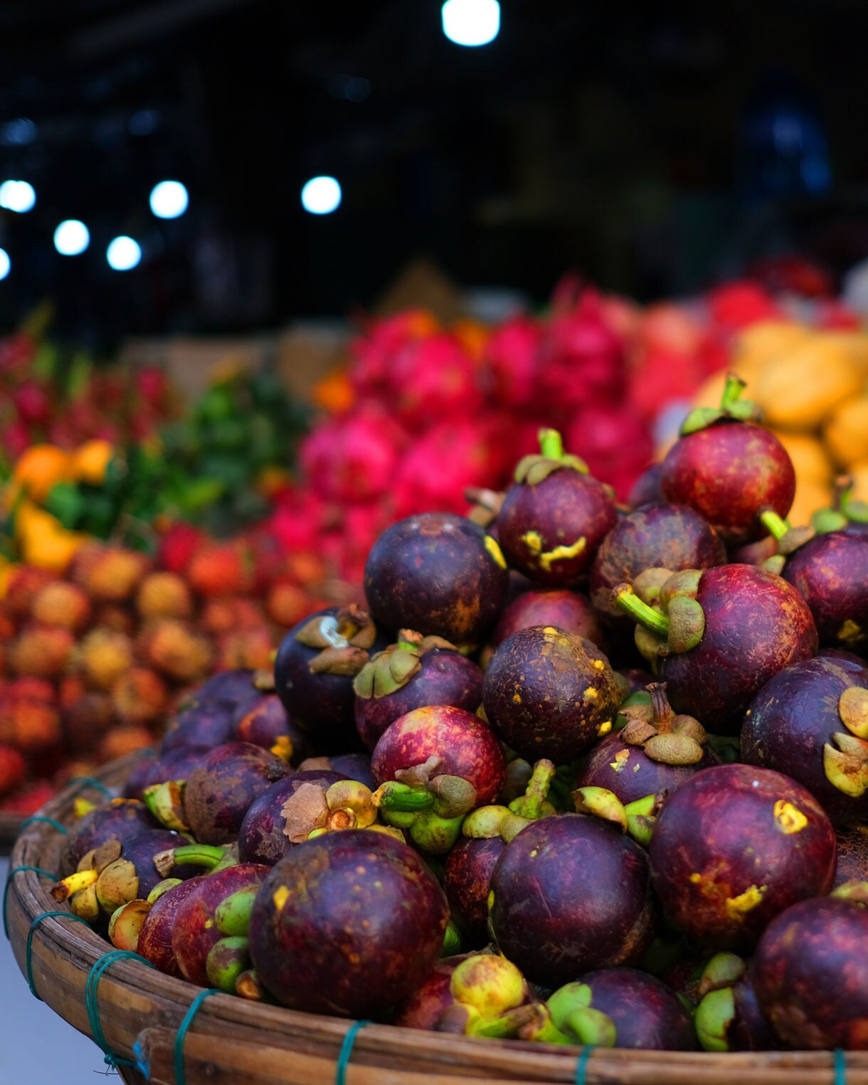Basket of ripe mangosteens at a Vietnamese fruit market, surrounded by colorful dragon fruit, mangoes and citrus in the background.