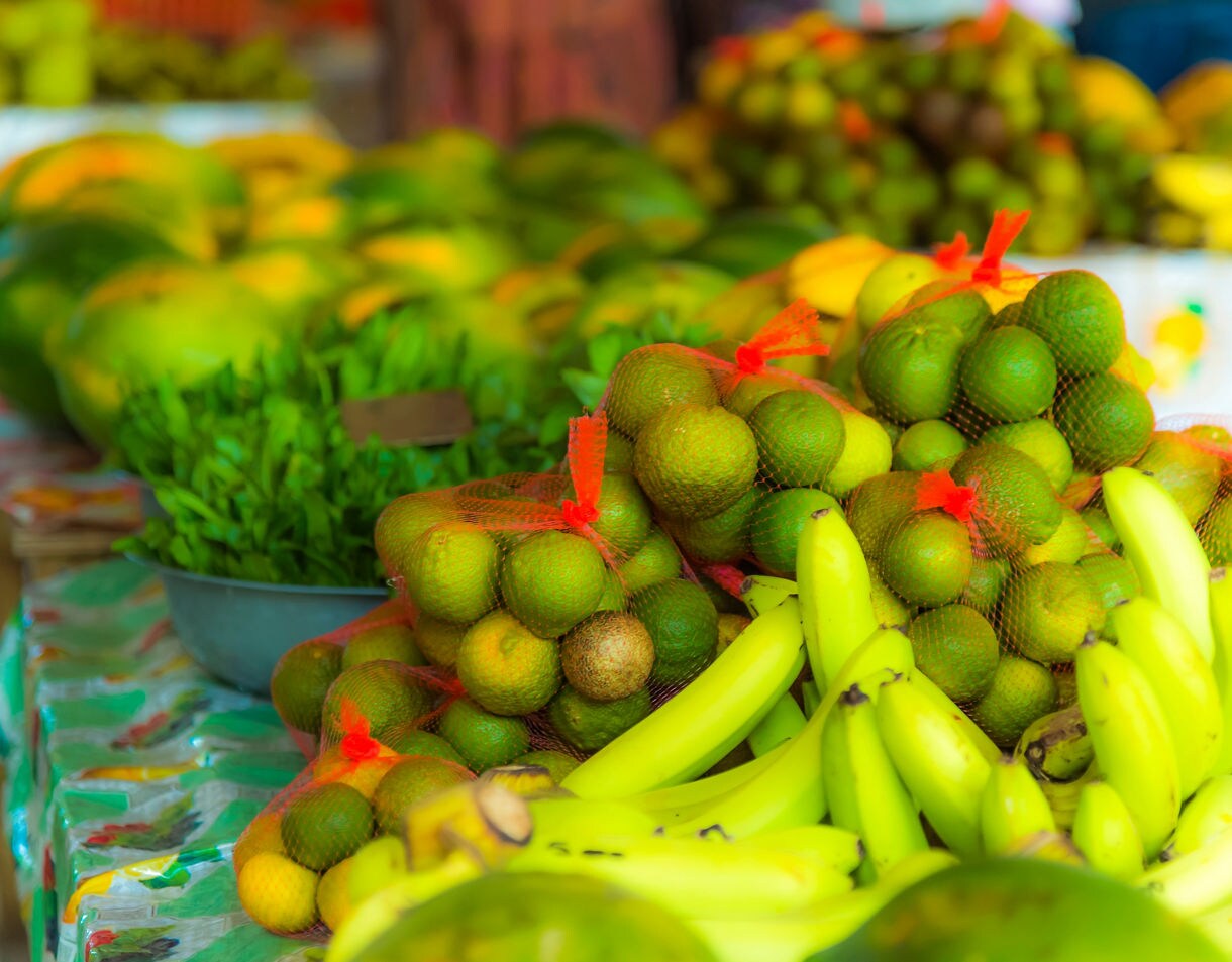 Close-up of bright green limes in mesh bags and yellow bananas on a market table, surrounded by assorted tropical produce.