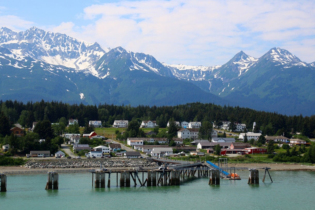 A scenic view of Fort William H. Seward from the Chilkoot Inlet in Haines, Alaska, featuring historic white buildings, lush greenery, and majestic snow-covered mountains in the background.