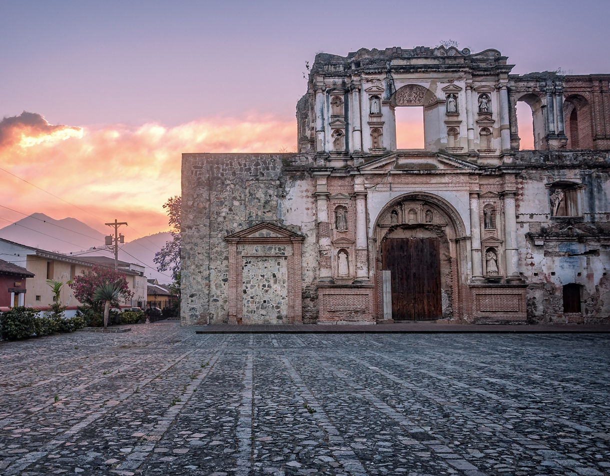 The ruins of a colonial church in Antigua, Guatemala, with the Fuego Volcano erupting in the distance under a pink and orange sunset sky.