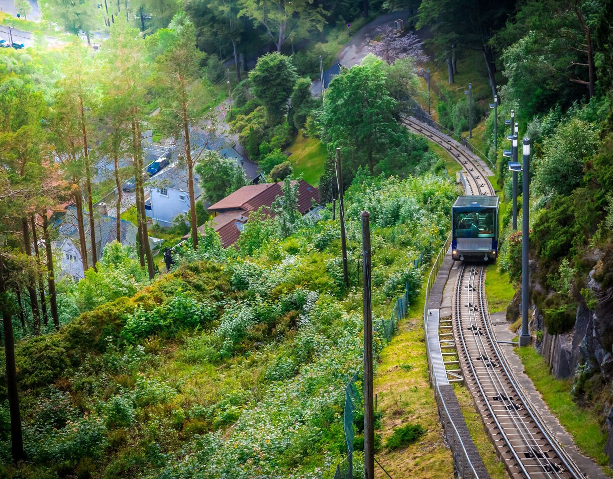 Glass-roofed Fløibanen funicular ascending through green forested slopes and rocky cliffs on Mount Fløyen in Bergen, Norway.