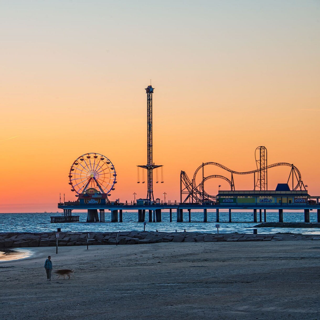 Sunset view of the Galveston Island Historic Pleasure Pier with amusement rides silhouetted against the orange and pink sky.