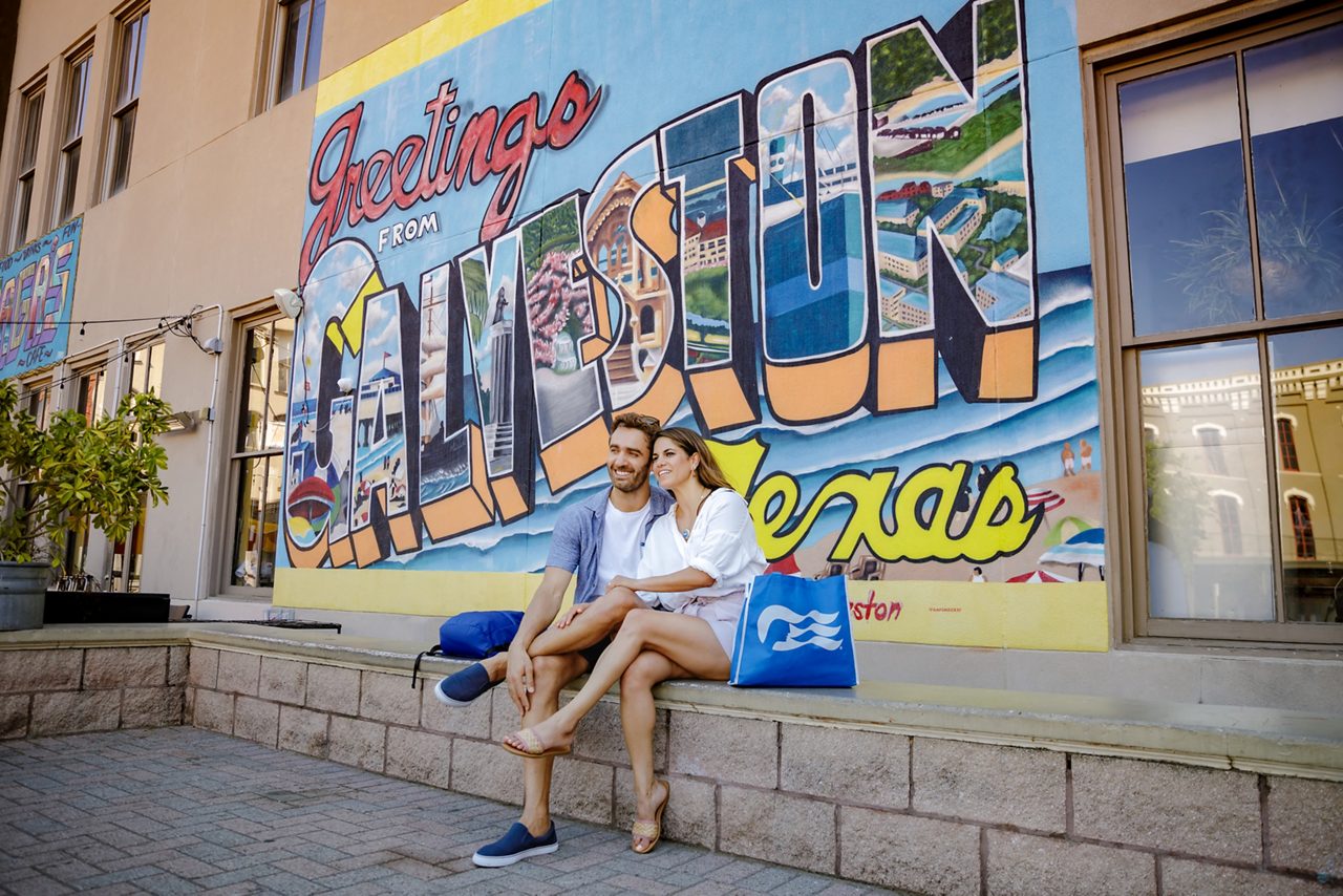 Picture a romantic couple standing in front of the iconic 'Greetings from Galveston' mural, holding Princess Cruises bags and smiling for a memorable photo.