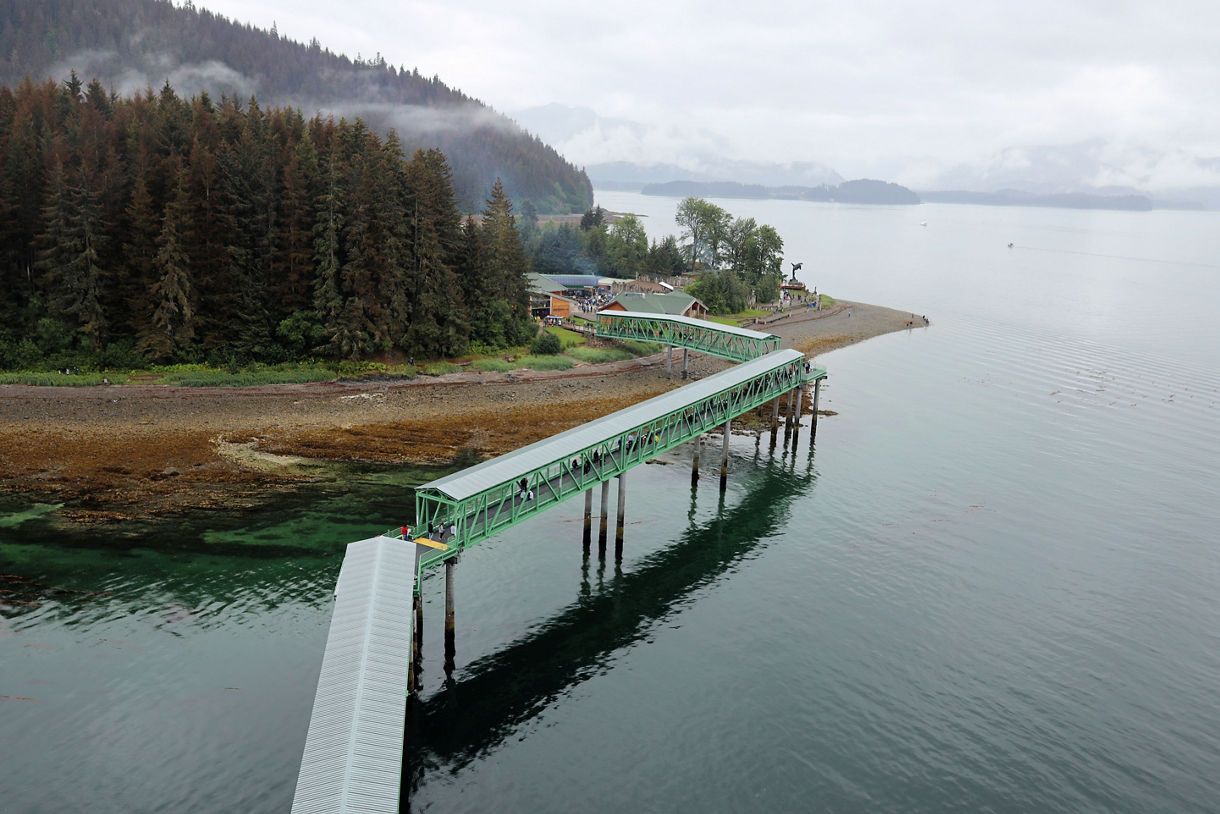 Icy Strait Point, Alaska, with the Ocean Landing Cruise Dock, green rampways, Adventure Center, orca sculpture, and nearby Hoonah village, surrounded by forest and ocean.