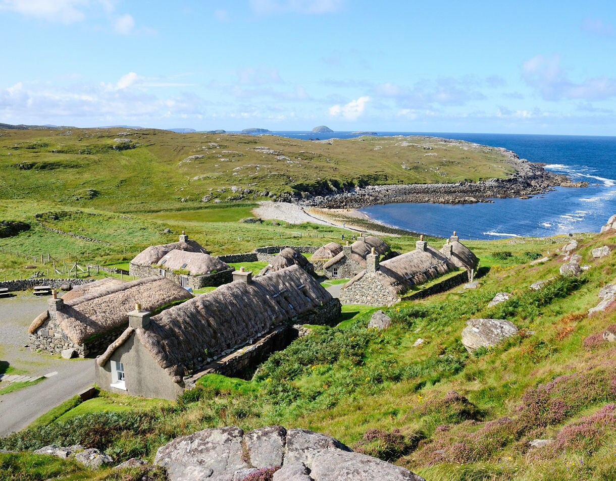 A scenic view of Gearrannan Blackhouse Village on the Isle of Lewis, featuring traditional stone cottages with thatched roofs set on rolling green hills overlooking a rocky shoreline and the blue Atlantic Ocean.