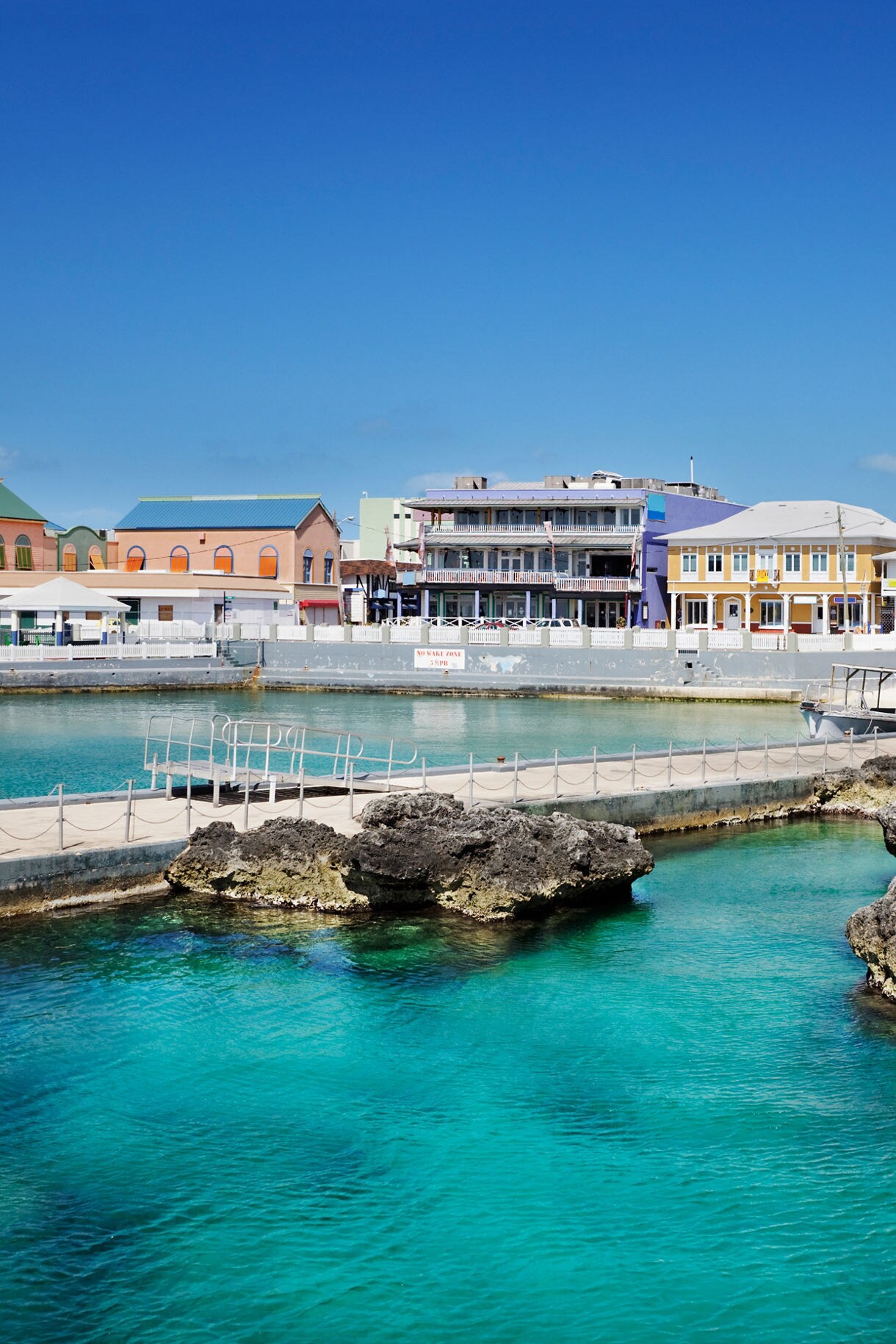 Colorful colonial buildings along George Town's waterfront in Grand Cayman with bright turquoise harbor waters and limestone formations in the foreground.