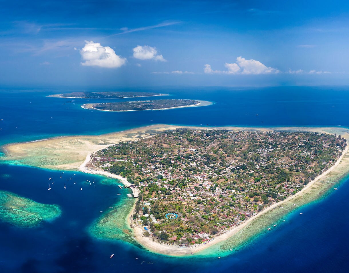 Aerial view of the Gili Islands showing a lush green island ringed by bright coral reefs and shallow turquoise water, with deeper blue ocean surrounding the coastline.
