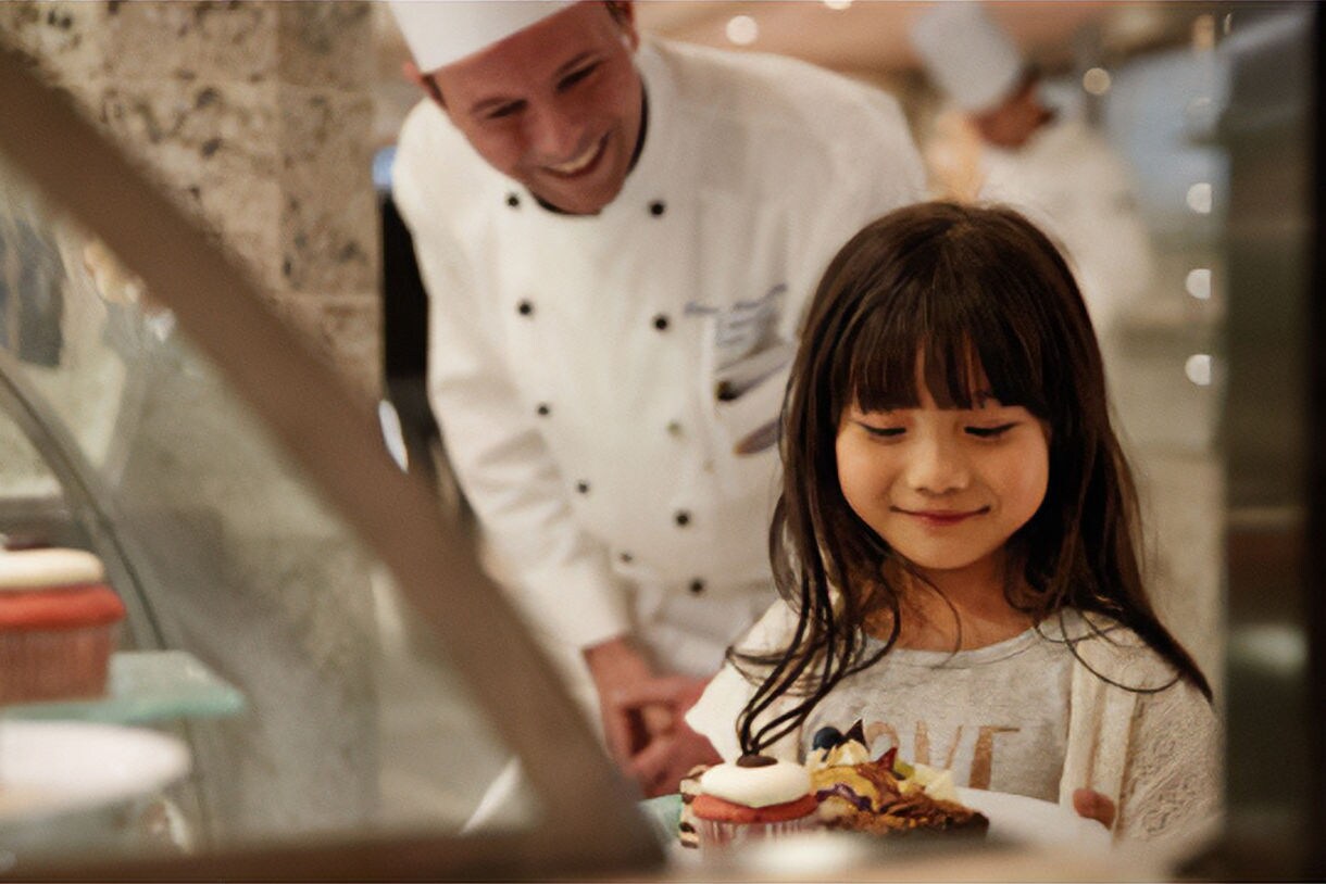 A smiling pastry chef helping a young girl select a treat from a glass display case filled with colorful desserts.