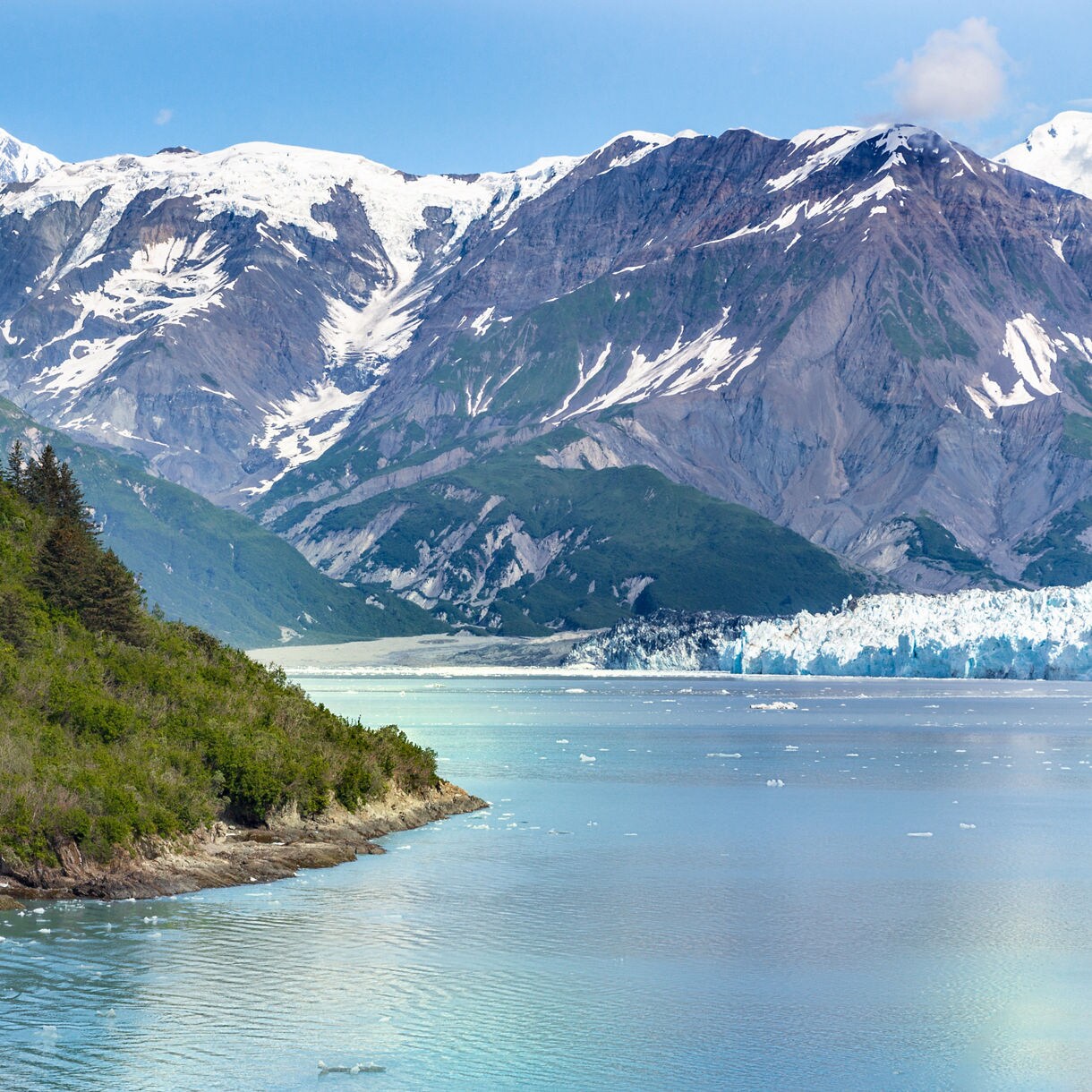 Scenic view of snow-covered mountains rising above turquoise glacial waters with green forested hills in the foreground near Juneau, Alaska.