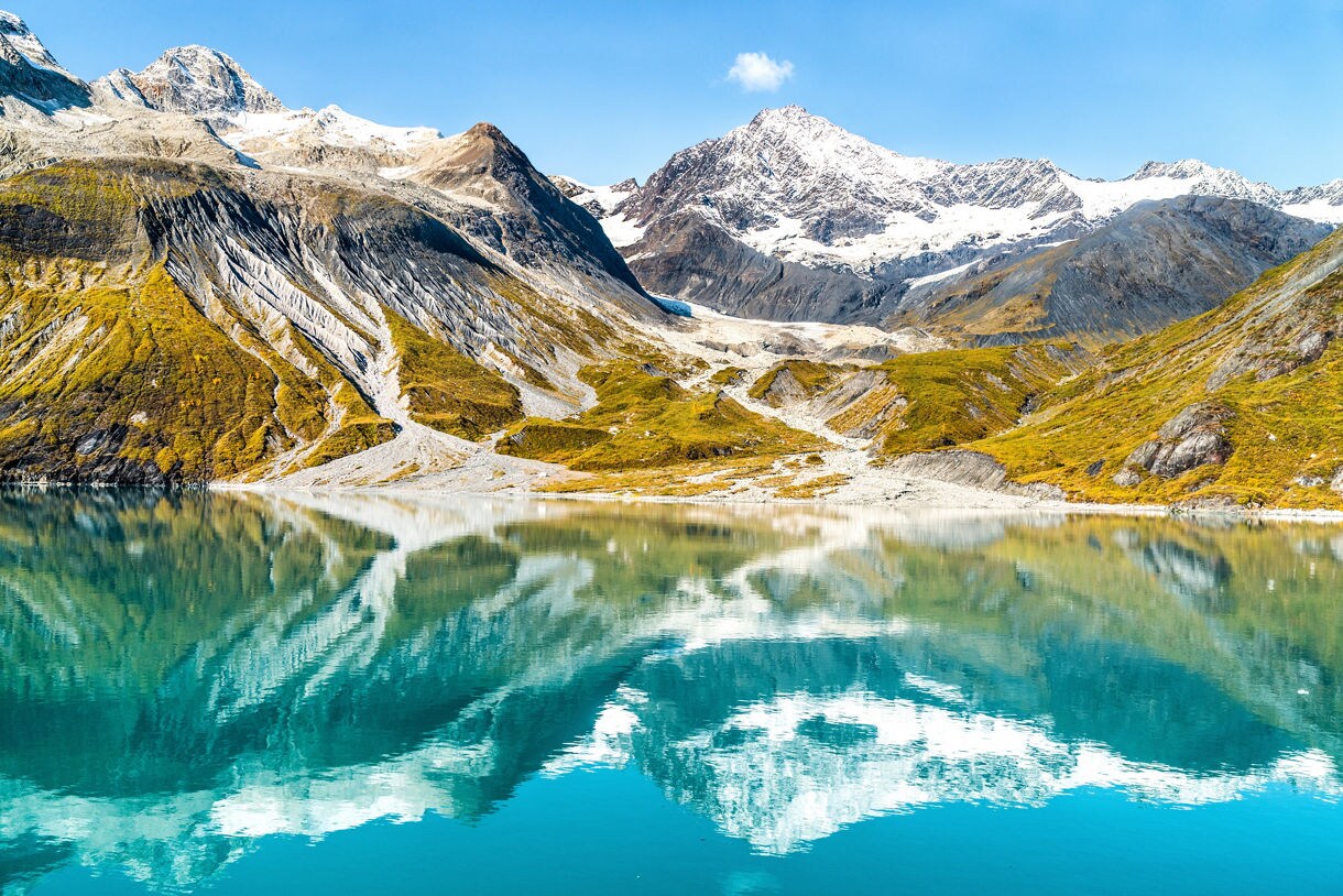 View of Glacier Bay National Park with snow-capped mountains and vibrant green slopes, their reflections shimmering in the calm turquoise waters below.