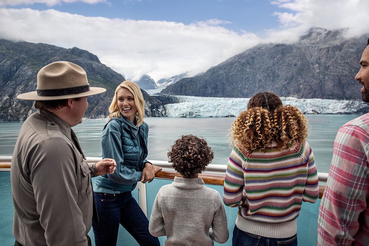A group of people stands on the deck of a cruise ship, gazing at a stunning glacier and mountain landscape. The setting is outdoors, with clear blue water and dramatic, snow-capped peaks in the background. The group includes adults and children, dressed in casual, warm clothing suitable for cool weather. The mood is relaxed and awe-inspired, with natural colors and soft lighting.