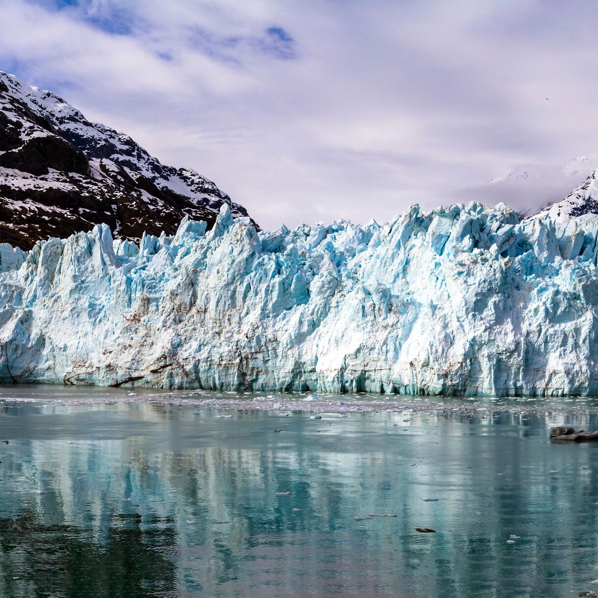 A view of Margerie Glacier in Glacier Bay National Park, with towering blue ice and snow-capped mountains reflecting in the still waters below.