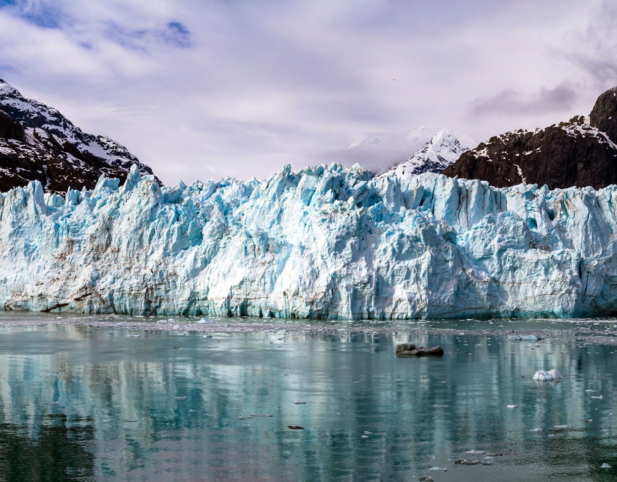 A view of Margerie Glacier in Glacier Bay National Park, with towering blue ice and snow-capped mountains reflecting in the still waters below.