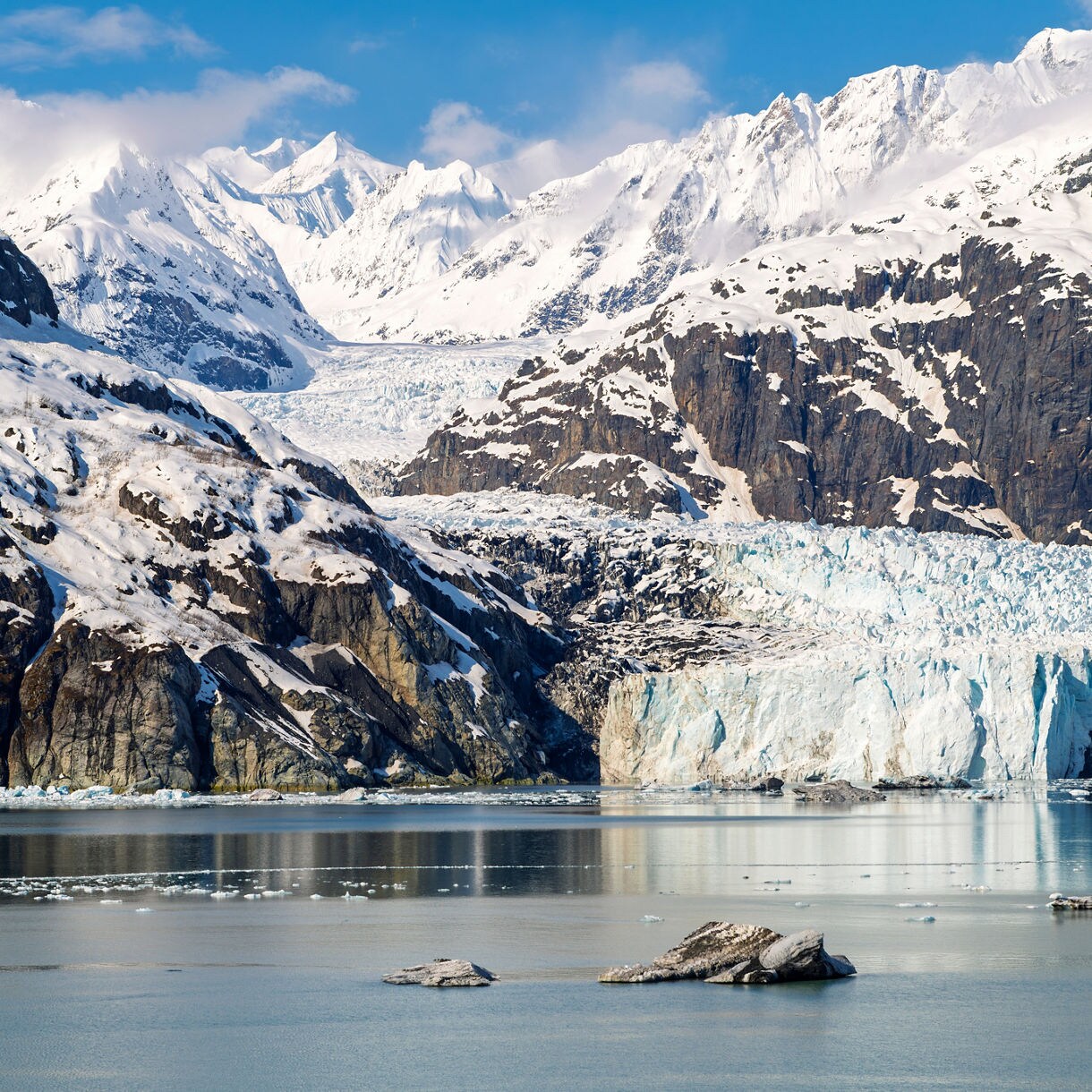 A scenic view of a majestic glacier flowing between snow-capped mountains, surrounded by a tranquil turquoise water in Glacier Bay National Park.
