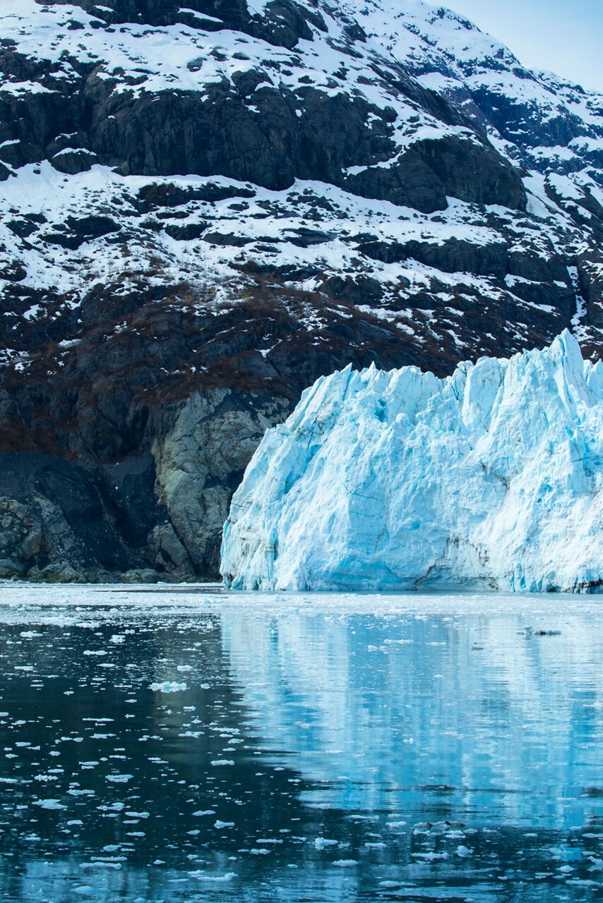 Close-up view of a towering glacier with deep blue ice, surrounded by snow-capped mountains and ice floes drifting in the calm water of Glacier Bay National Park.