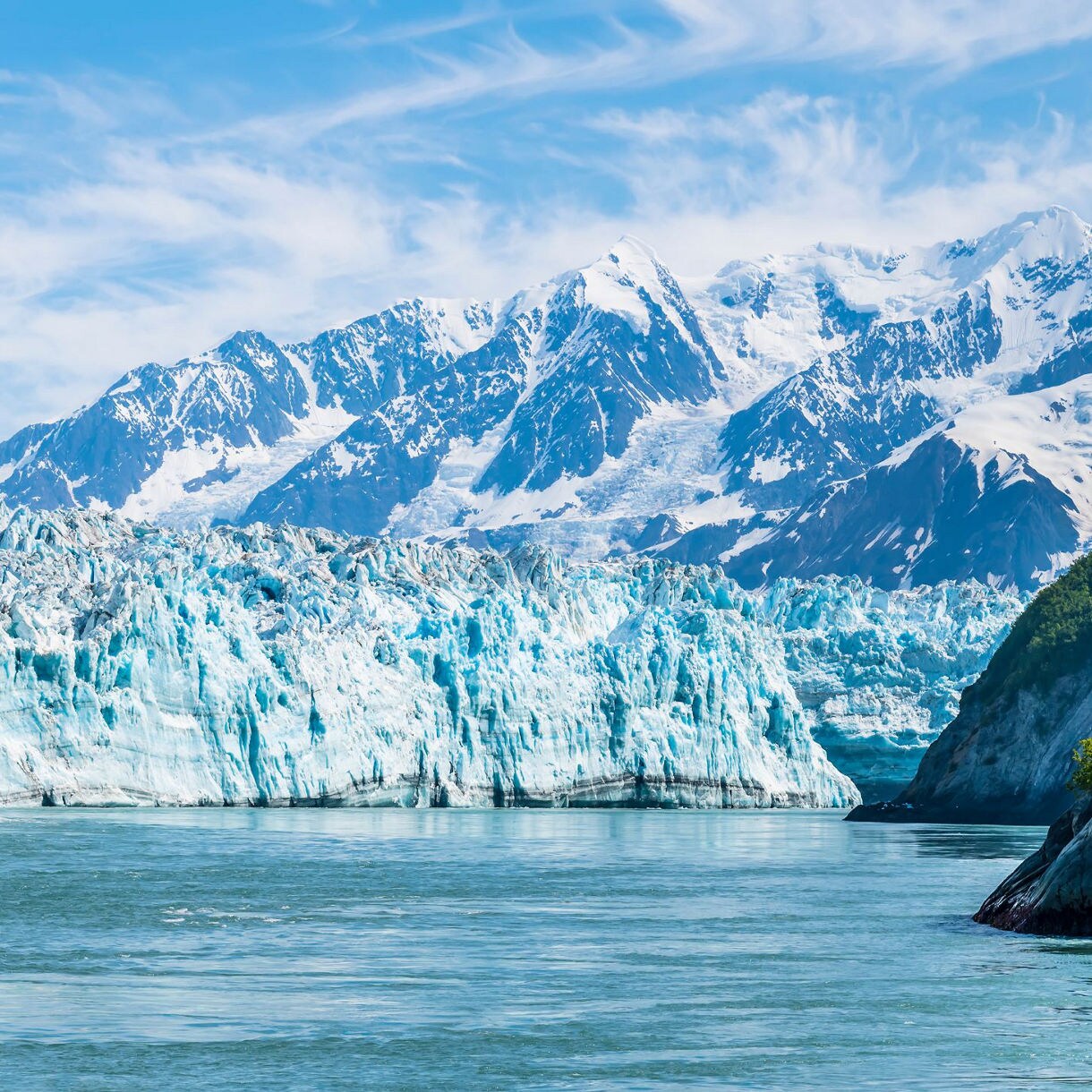 Large white and blue glacier flowing from snow-capped mountains into calm blue water with forested rocky shoreline at Glacier Bay, Alaska.