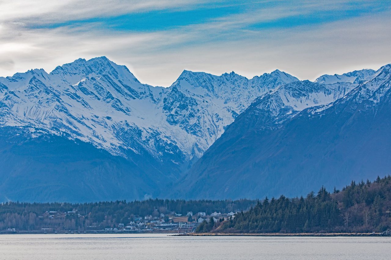 Snow-covered mountain range with forested foothills and small coastal community visible along waterfront in Haines, Alaska.