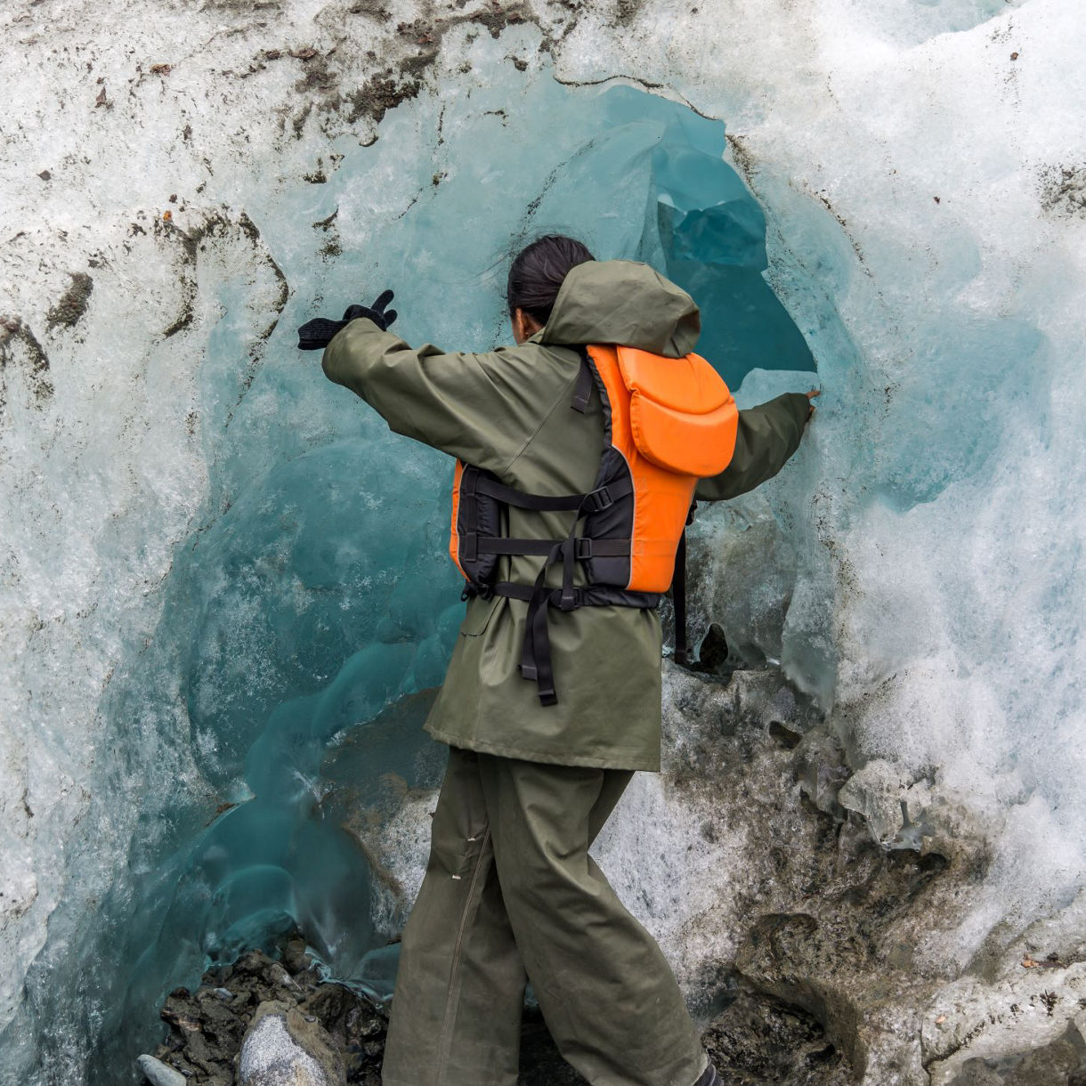 A tour participant in olive green field gear and an orange safety vest kneels beside a bright blue meltwater pool on a Skagway glacier tour, examining the pristine glacial ice formations.