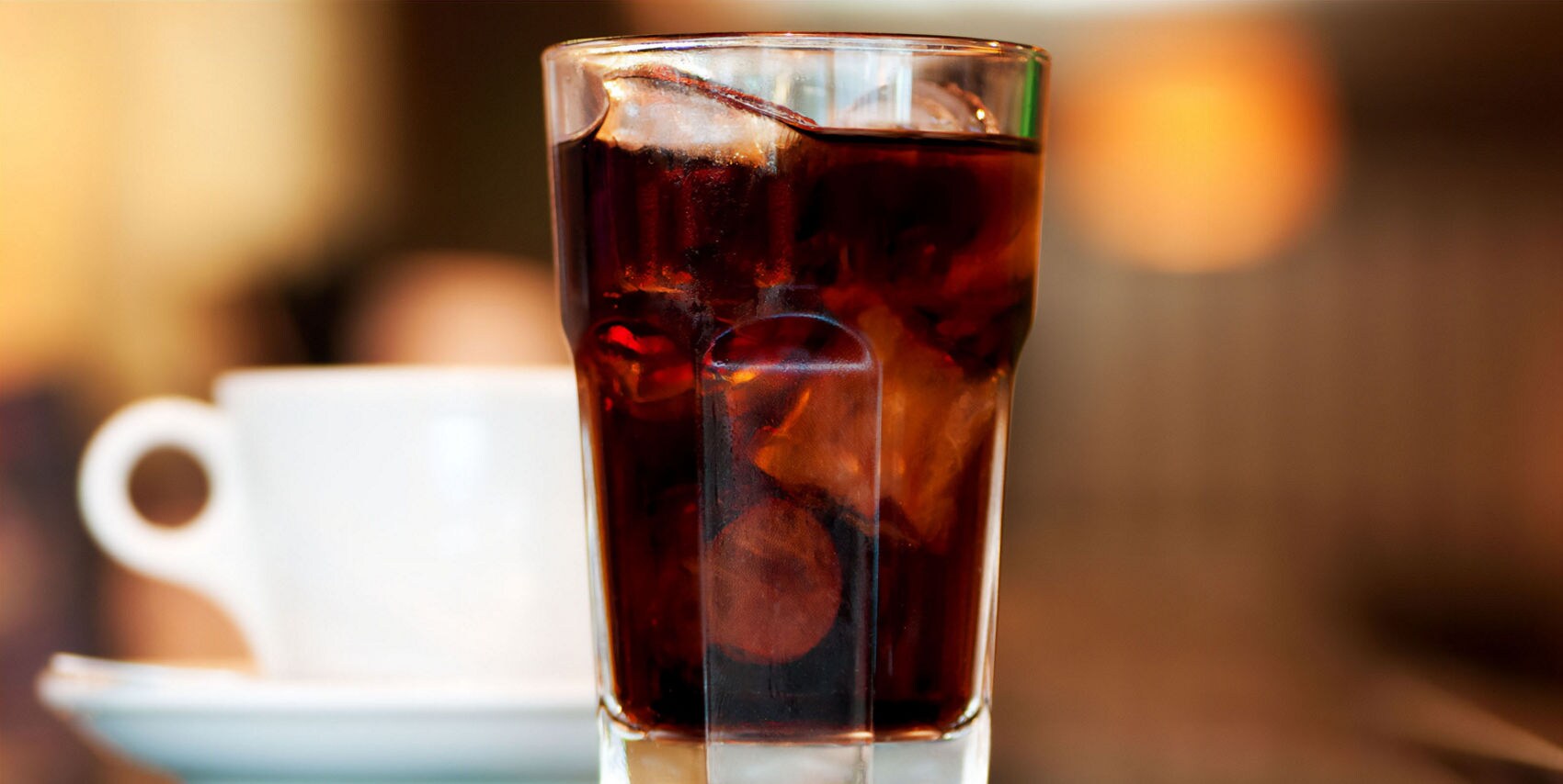 A tall glass of iced cola beside a white coffee cup on a saucer, set against a warm, blurred background.
