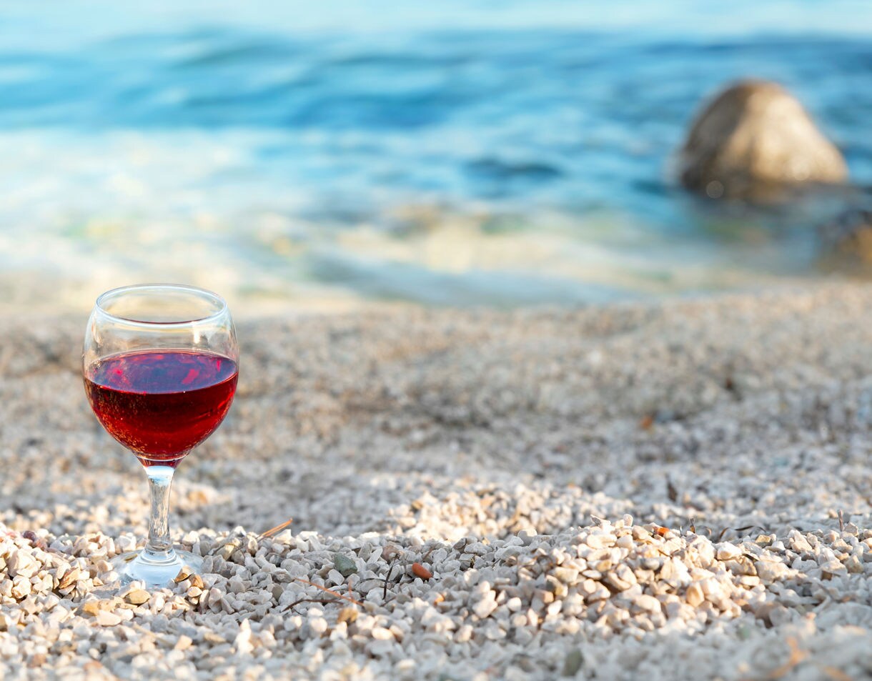 A glass of red wine resting on a pebbled beach with soft blue waves in the background.