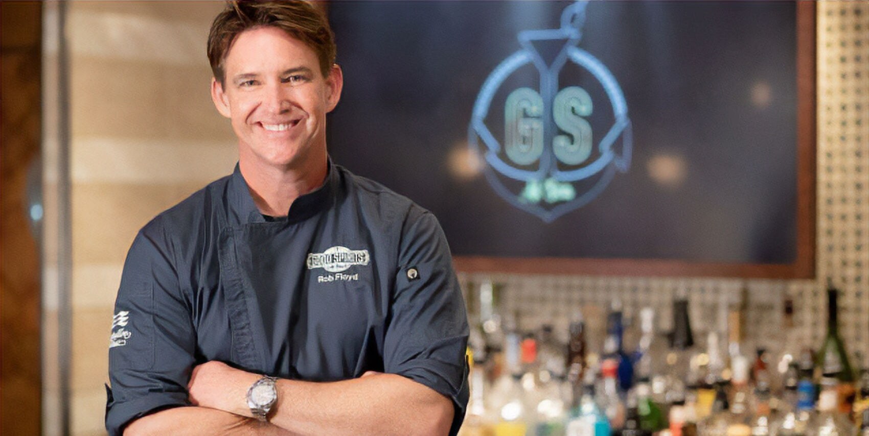Rob Floyd in a dark chef's jacket stands with arms crossed in front of the glowing Good Spirits At Sea logo.