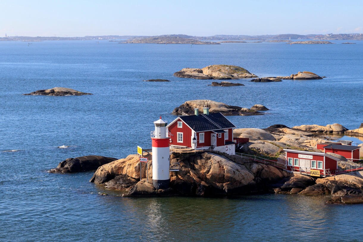 A small, rocky island features a red wooden cottage and a red-and-white lighthouse surrounded by blue sea waters under a clear sky.
