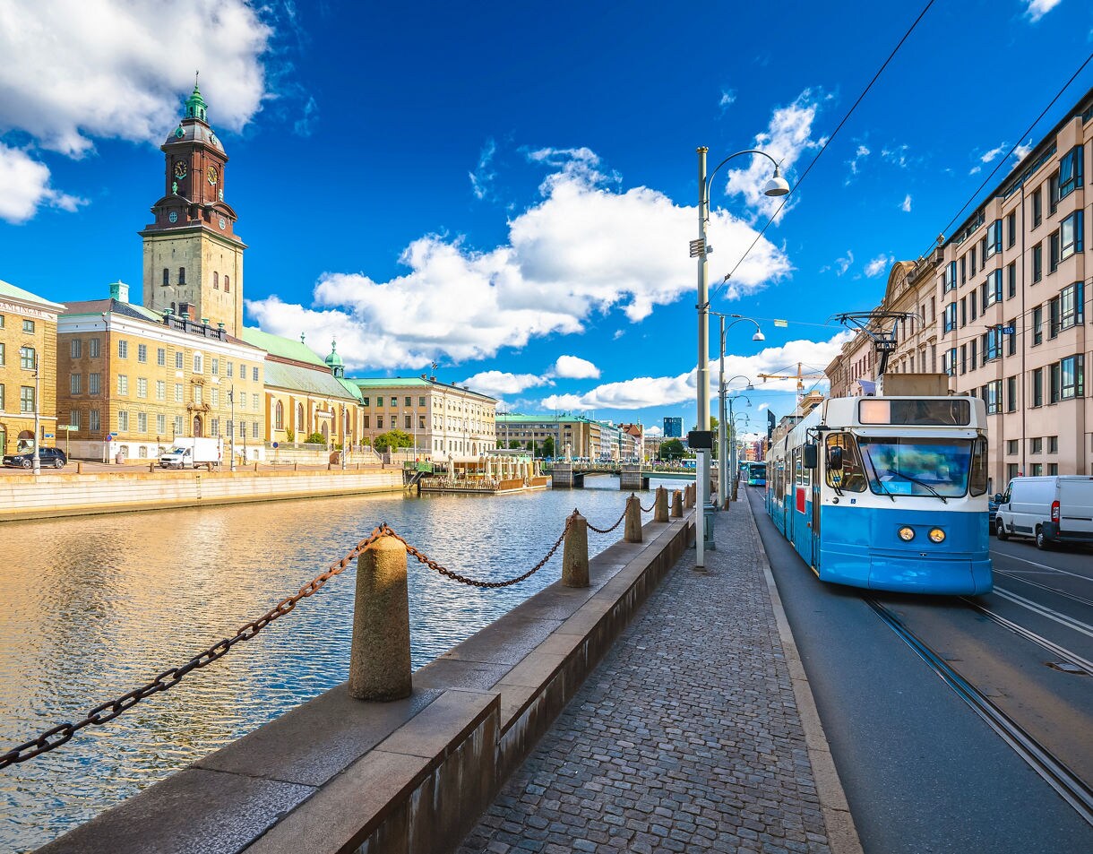 A classic blue-and-white Gothenburg tram travels along a street parallel to a canal lined with stone bollards, historic yellow-brick buildings, and the prominent tower of the German Church under a bright blue sky with fluffy white clouds.