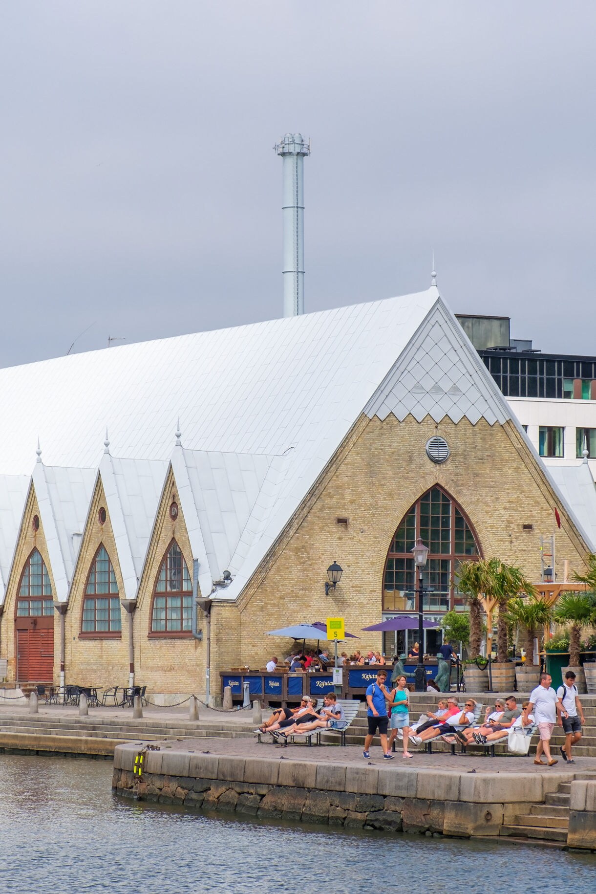 A distinctive historic market hall with a steep white roof and pointed arch windows stands along a calm waterfront. People walk and relax on the quay in front of the building, adding a lively urban atmosphere. Modern office buildings and industrial chimneys rise in the background, contrasting with the traditional architecture. The scene is captured on a bright, slightly overcast day, highlighting the mix of heritage and contemporary city life..