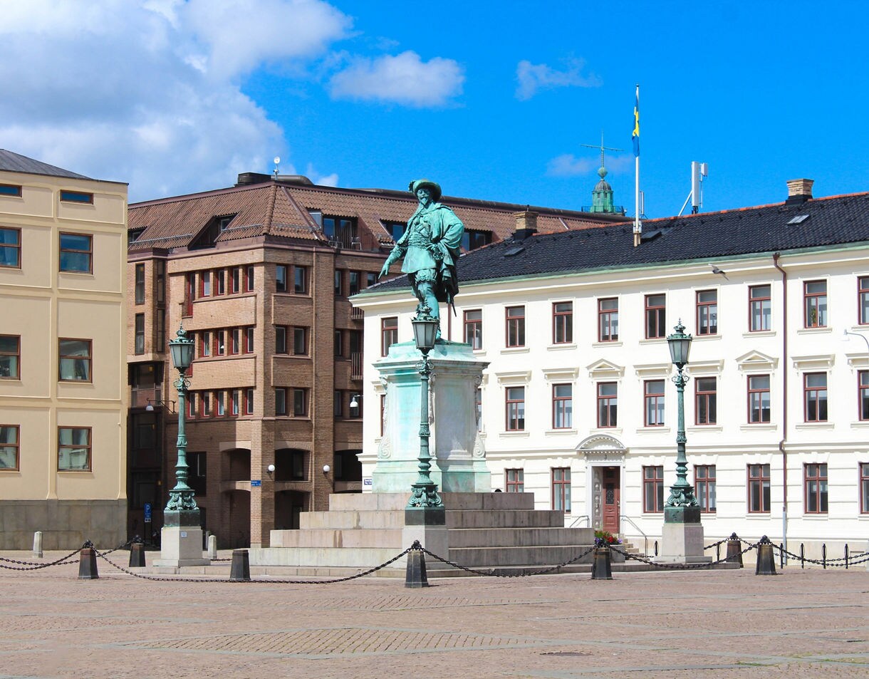 A bronze statue of King Gustav II Adolf stands atop a grand stone pedestal in the center of an open plaza, flanked by ornate lampposts and classic European architecture under a bright blue sky.