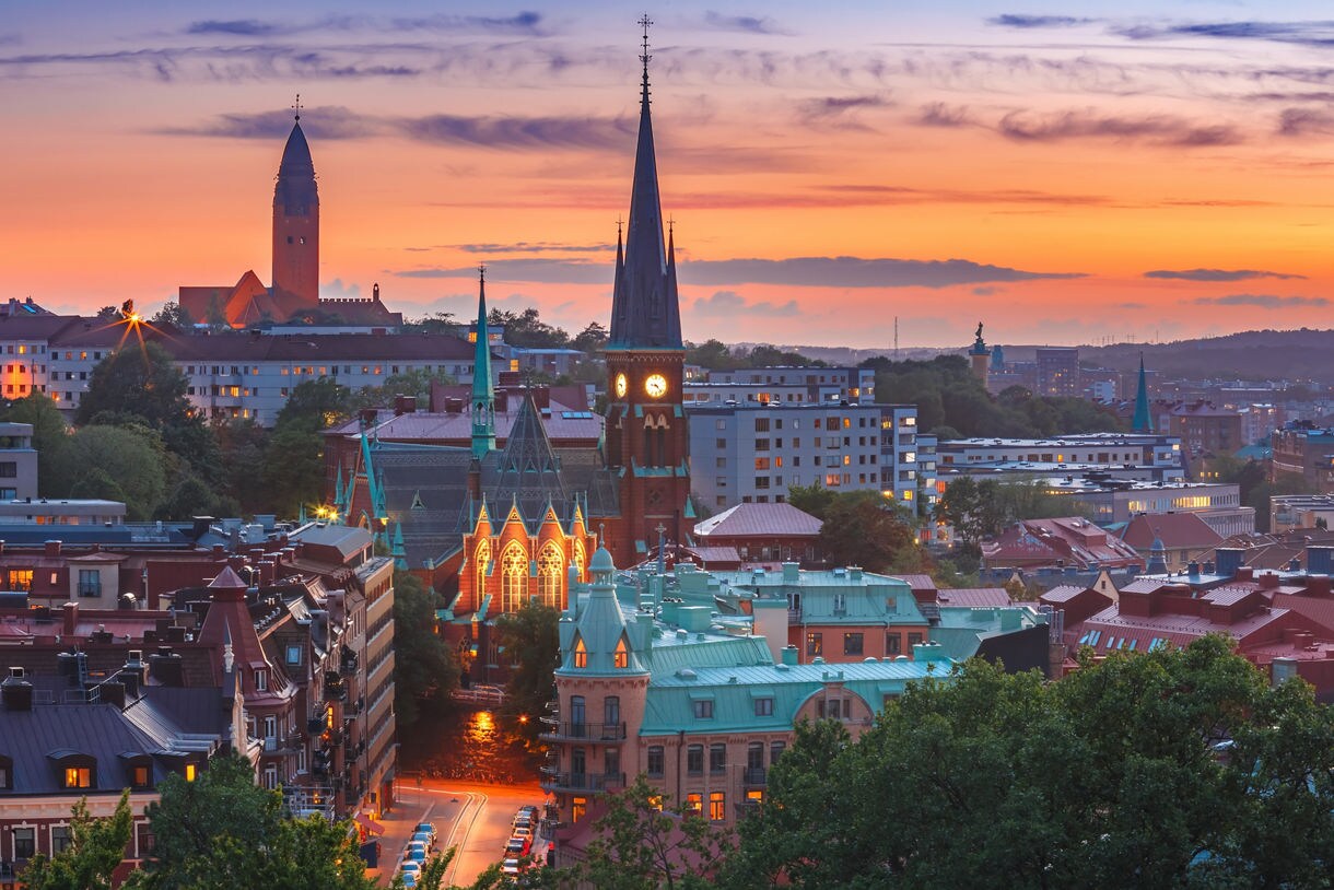 A high-angle city view shows the illuminated Oscar Fredrik Church and surrounding green-roofed buildings at dusk beneath a vibrant orange and purple sunset sky.