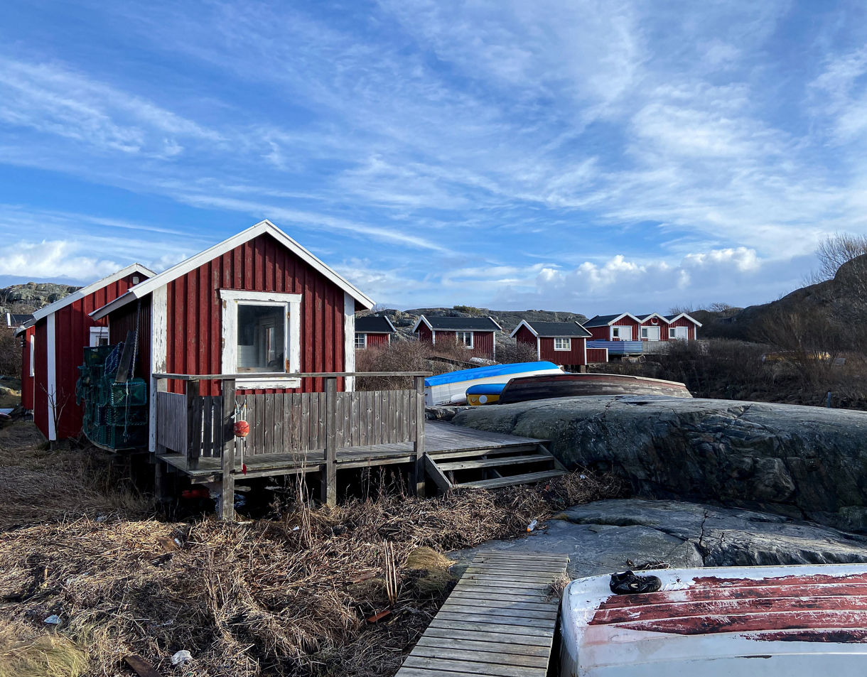 Red wooden fishing huts with white trim sit on a rocky shoreline with wooden decks, boardwalks, and small overturned boats under a partly cloudy blue sky.