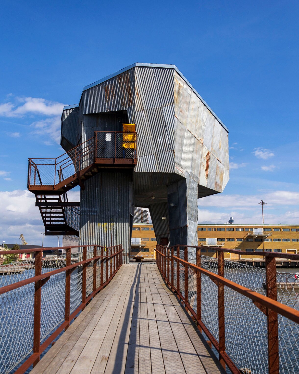 A wooden walkway with rusty metal railings leads toward a unique, elevated sauna made of corrugated metal cladding set over water under a clear blue sky.