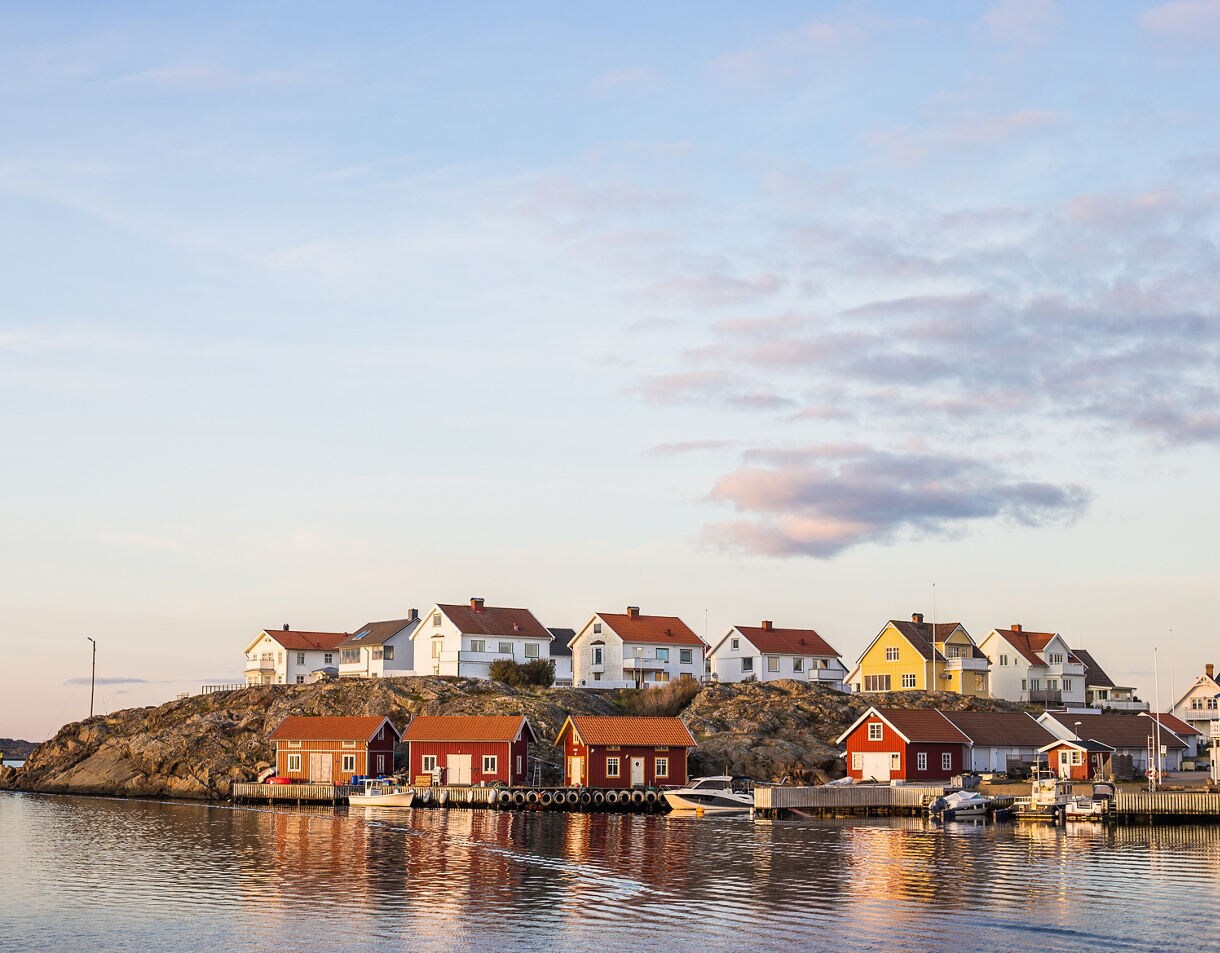 Red boathouses with wooden docks sit along the water's edge below a rocky hill lined with white and yellow houses under a soft, twilight sky.