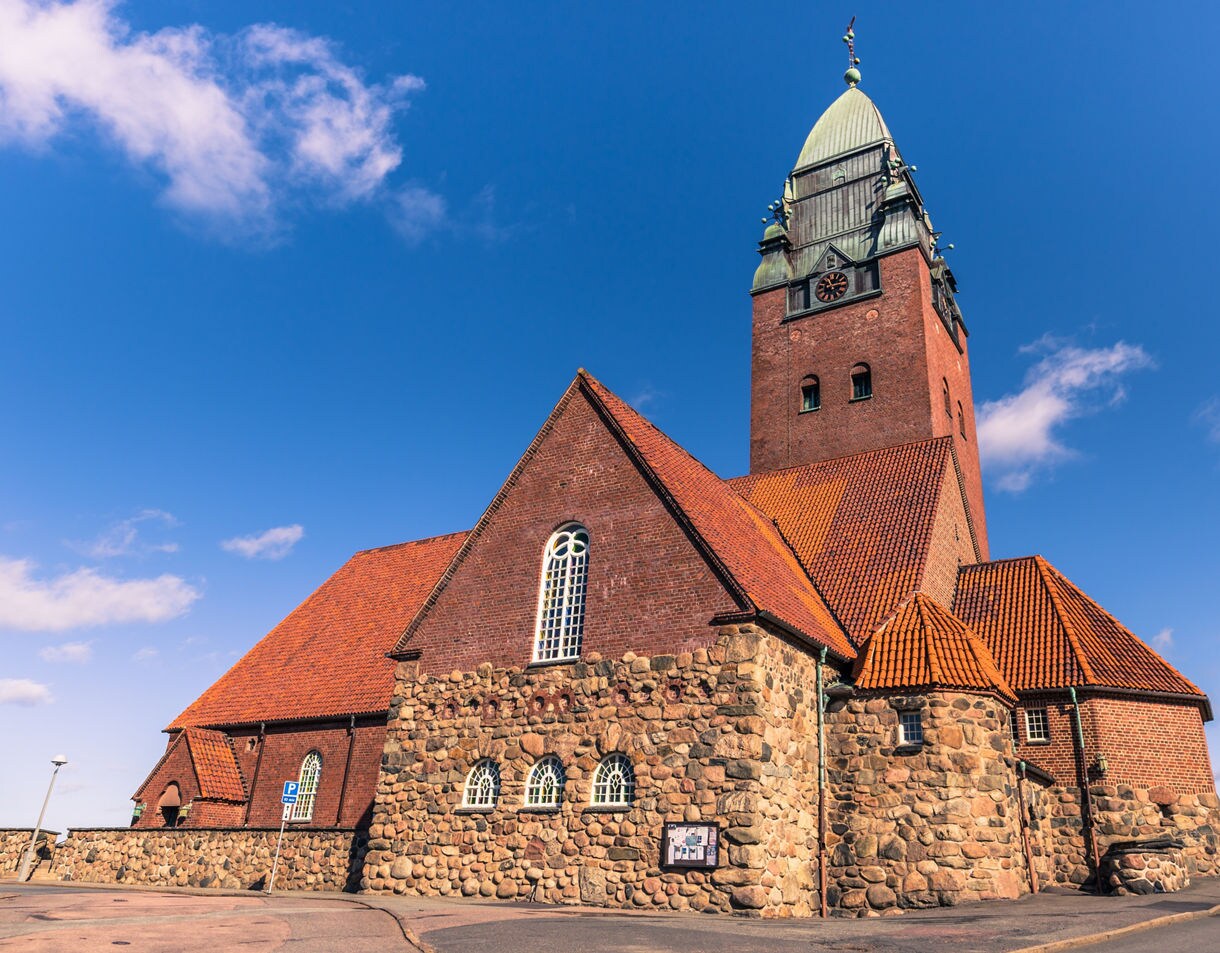 A large red-brick and fieldstone church features a red-tiled roof and a tall, distinctive clock tower with a green roof under a clear blue sky.