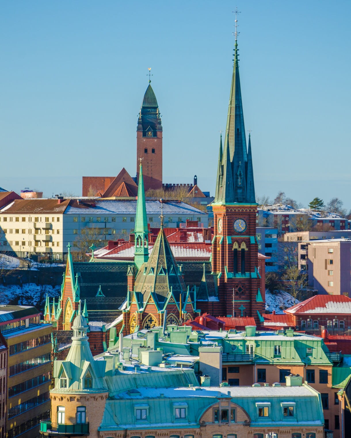 A vibrant cityscape features a prominent historic church with tall green spires and a red brick clock tower. Surrounding the church are residential and commercial buildings with varied architectural styles and colorful facades. Snow lightly covers rooftops, suggesting a cold winter day under clear blue skies. The composition highlights the contrast between traditional architecture and modern urban structures.