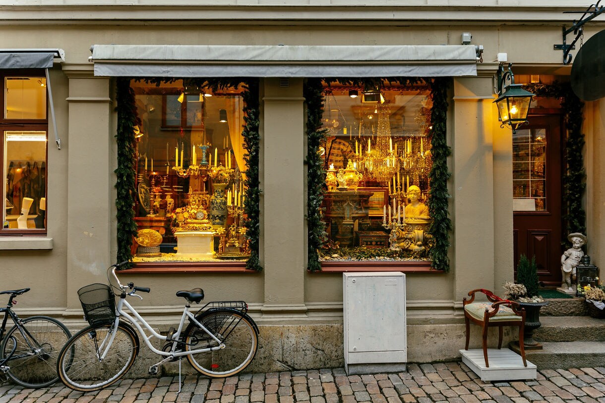 A white bicycle stands parked on a cobblestone street in front of a warm, illuminated antique shop window display framed with green garland.