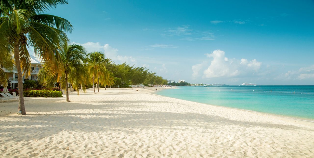 A serene view of Grand Cayman's Seven Mile Beach, with powdery white sand, swaying palm trees and turquoise waters stretching to the horizon.
