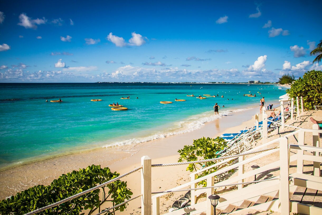 A vibrant view of Seven Mile Beach in Grand Cayman, featuring turquoise waters, golden sand and visitors enjoying the gentle waves under a bright blue sky.