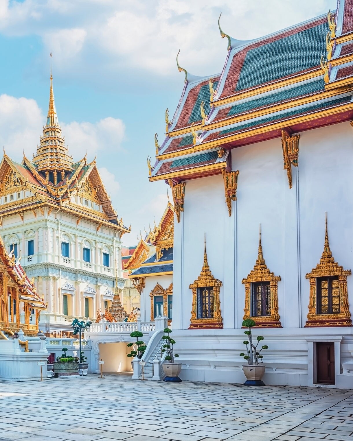 Intricate temple buildings of the Grand Palace in Bangkok, featuring gilded roofs, white walls and tall pointed spires under a bright sky.