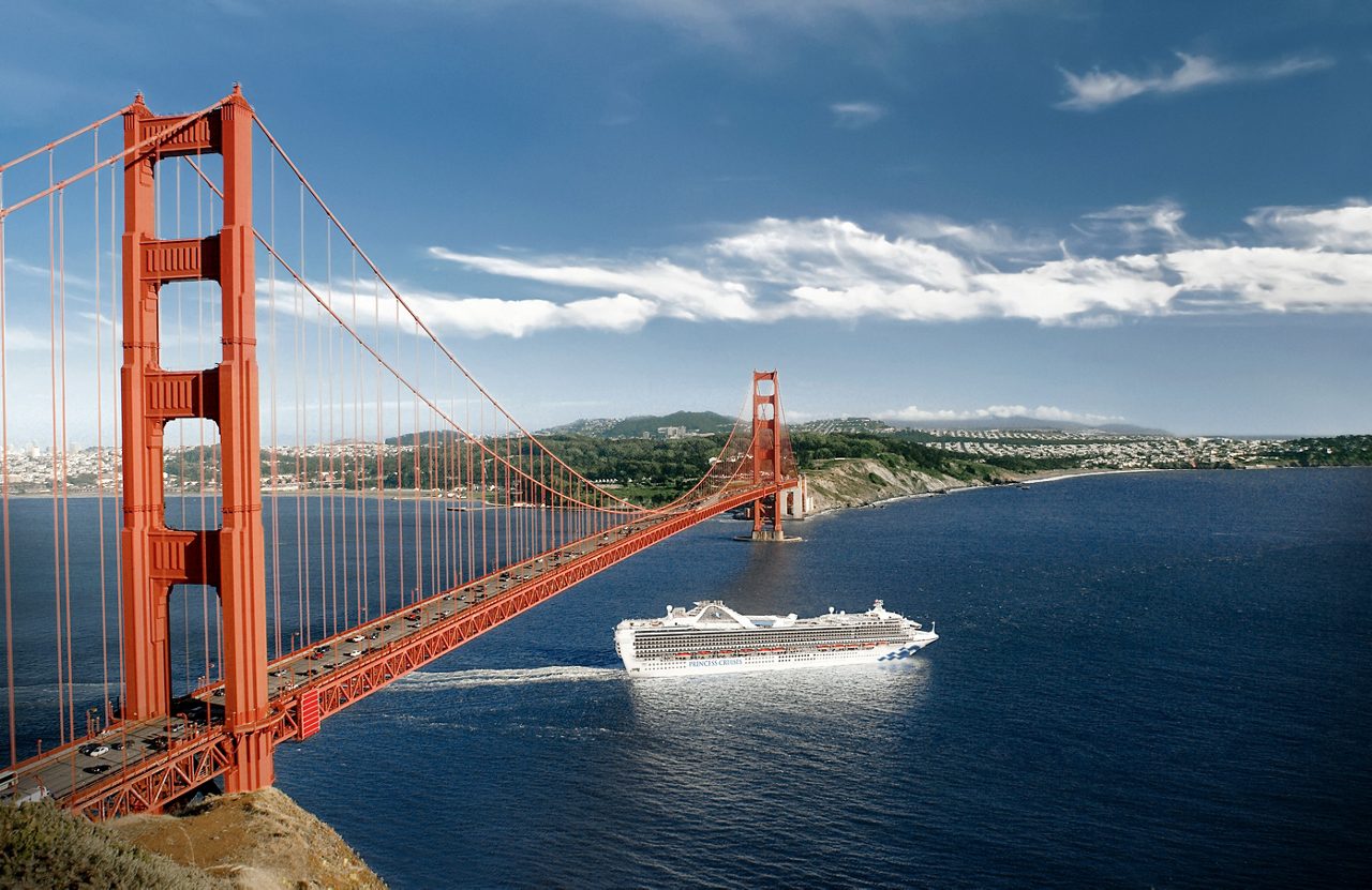 A large Princess cruise ship passing under a red suspension bridge over a body of water. 