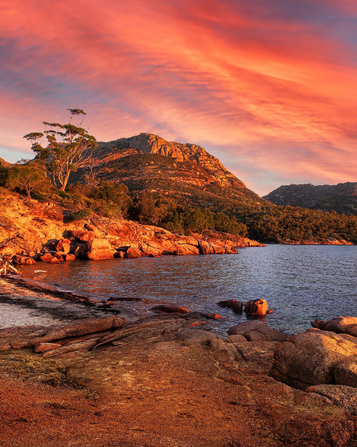 Rocky shoreline in Freycinet National Park at sunset, with the Hazards mountain range lit in warm orange light and dramatic red clouds stretching across the sky above calm coastal waters.