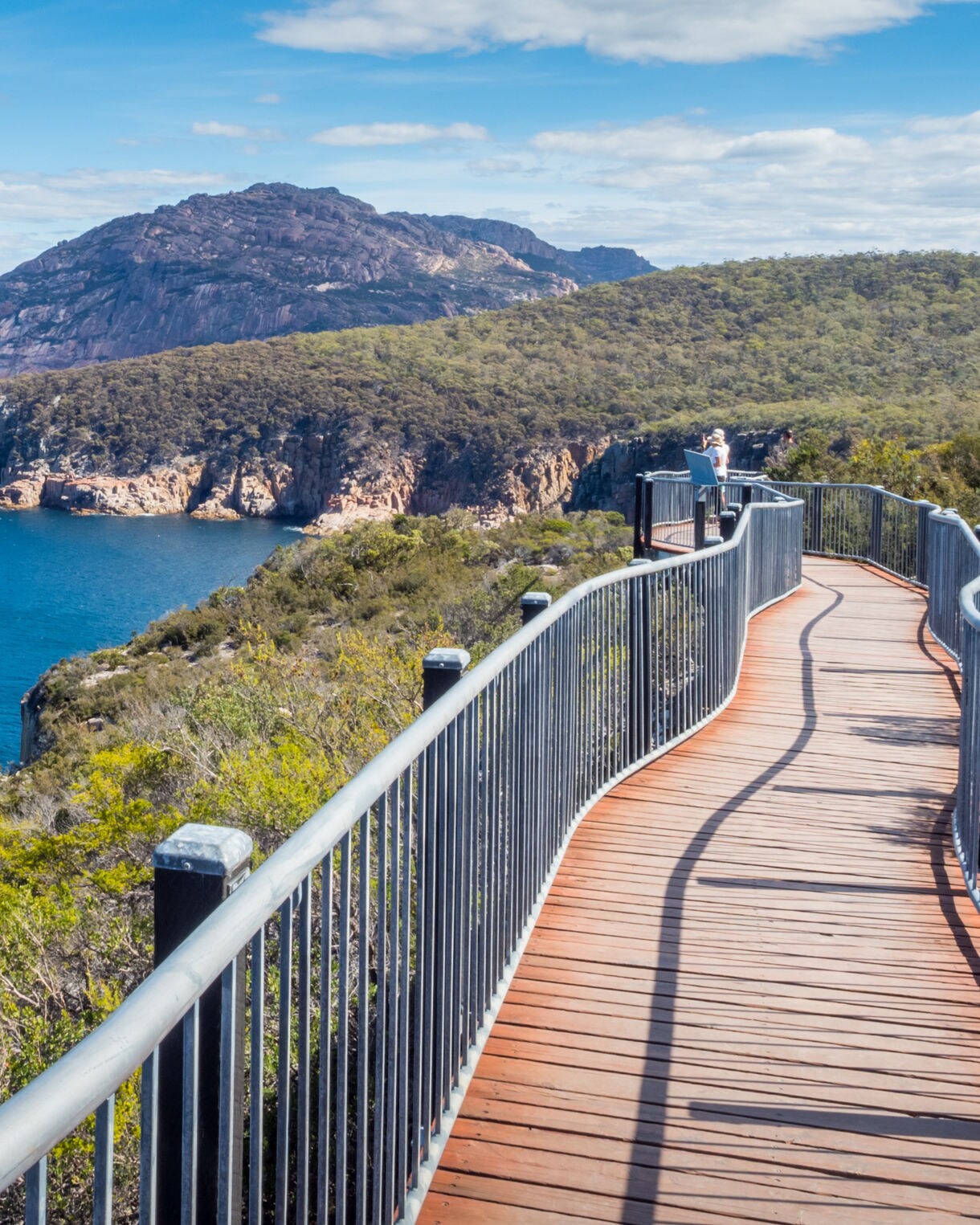 Curved boardwalk overlooking the rocky coastline and deep blue ocean in Freycinet National Park, with forested hills and mountains in the distance under a bright sky.