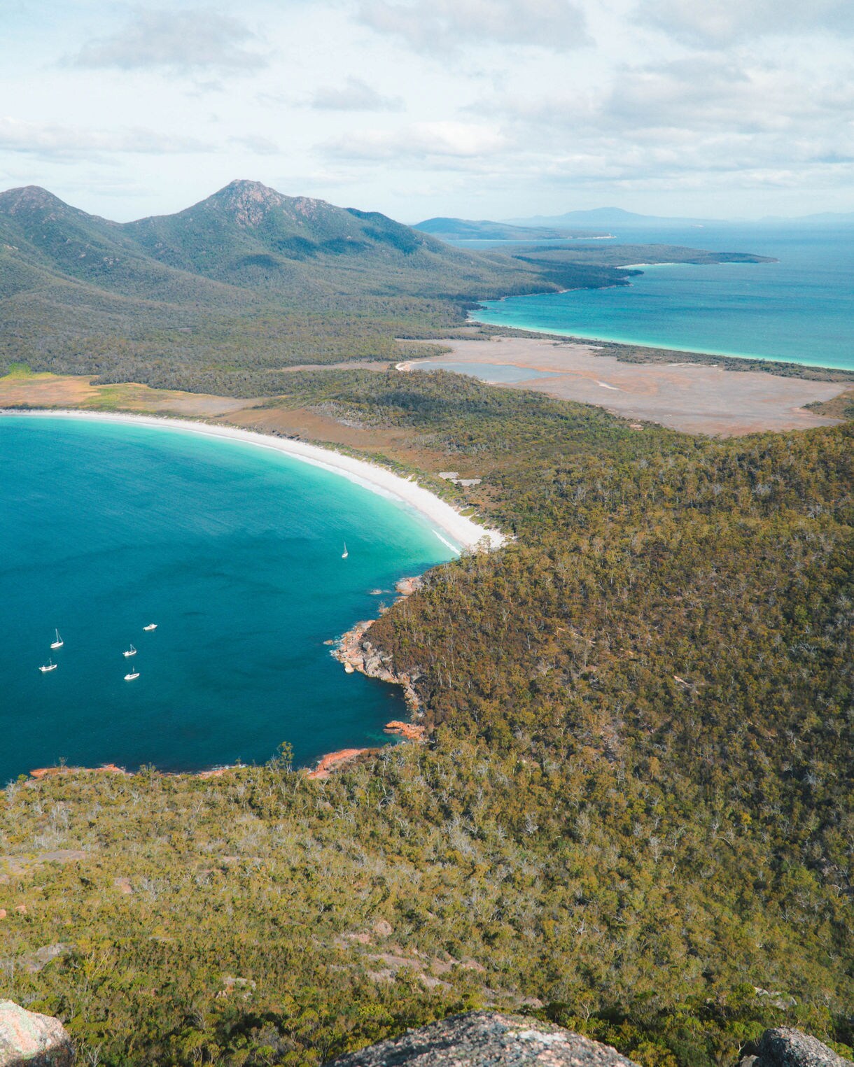 Vertical aerial view of Wineglass Bay in Freycinet National Park, showing a crescent of white sand, turquoise water with small boats, and forested mountains stretching into the distance.