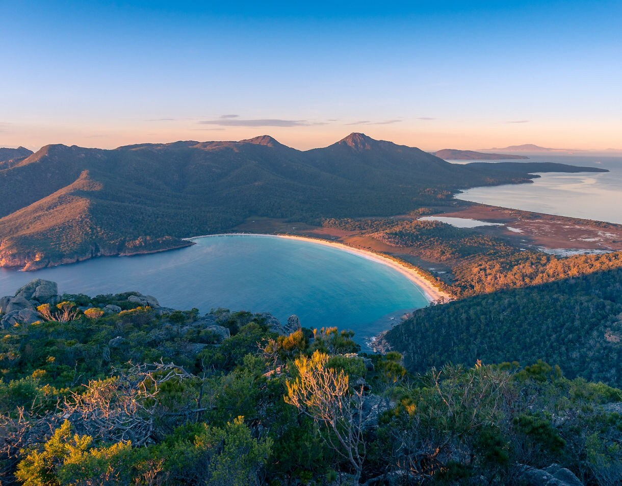 Elevated view of Wineglass Bay in Freycinet National Park at sunrise, showing a curved white-sand beach, turquoise water, forested mountains, and warm golden light illuminating the coastline.