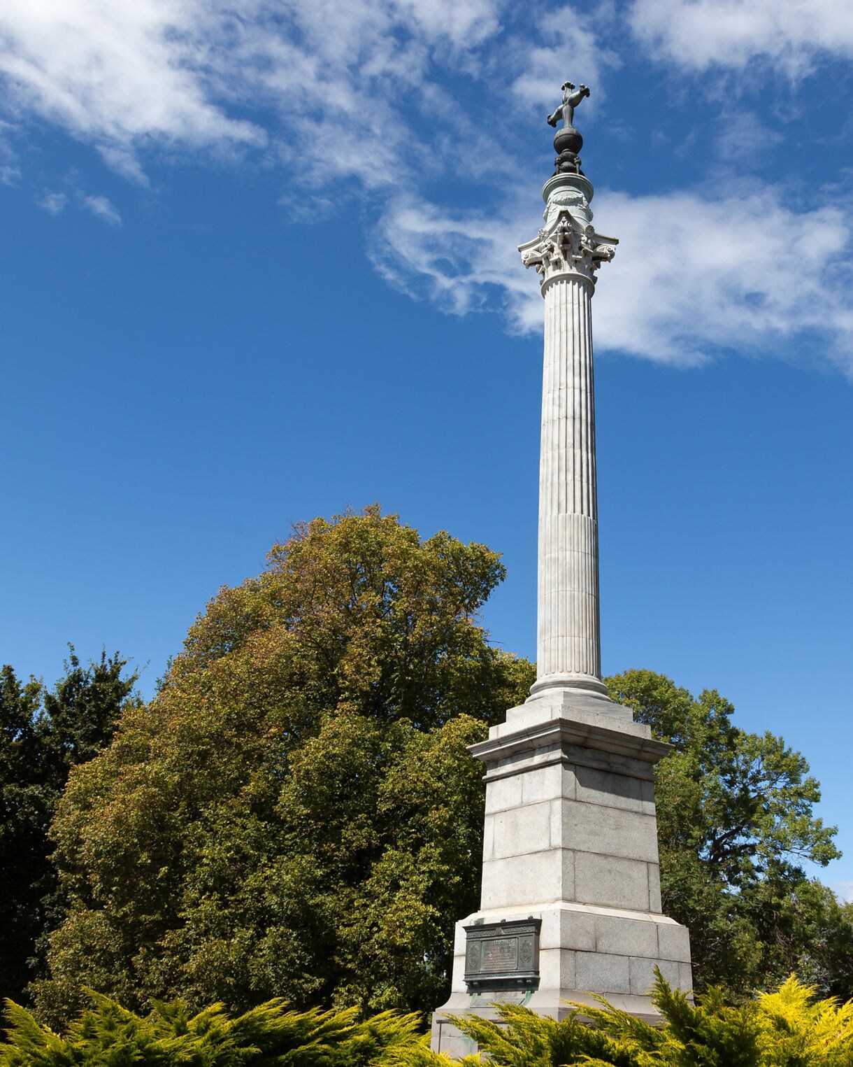 Tall stone war memorial column topped with a bronze figure, surrounded by green trees and flowering shrubs under a clear sky.