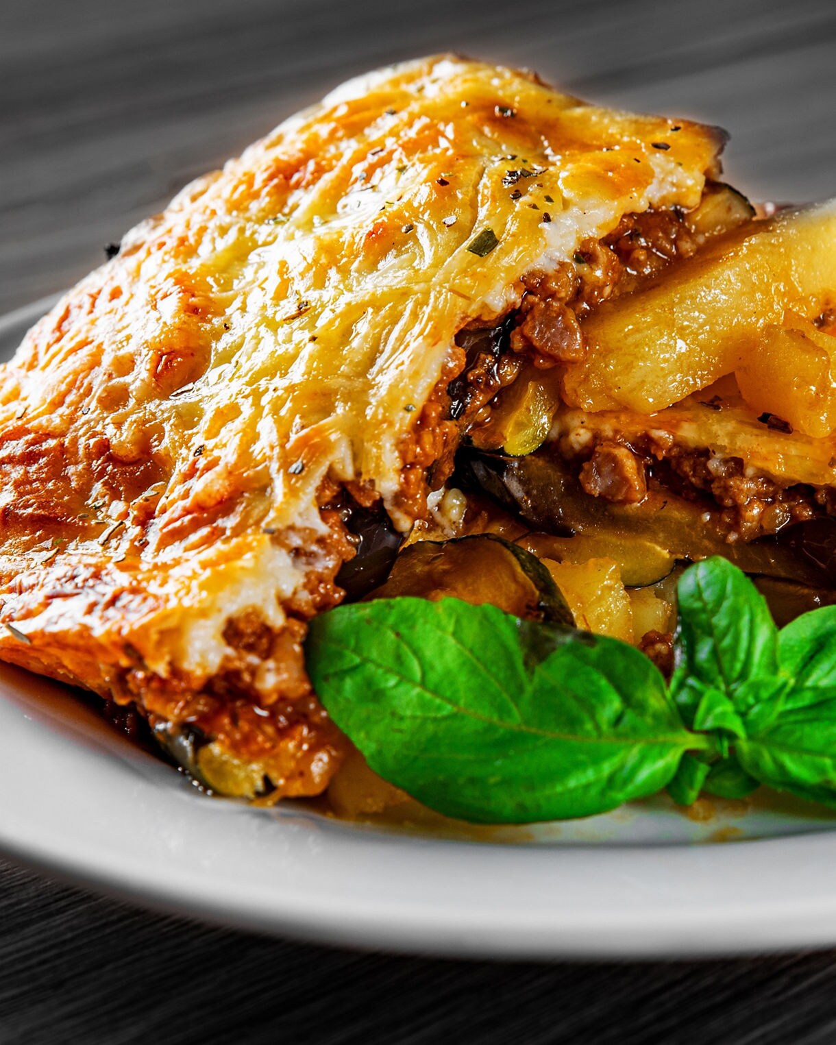 Close-up of a slice of baked Greek moussaka on a white plate, showing layers of eggplant, potatoes, meat sauce and golden béchamel.