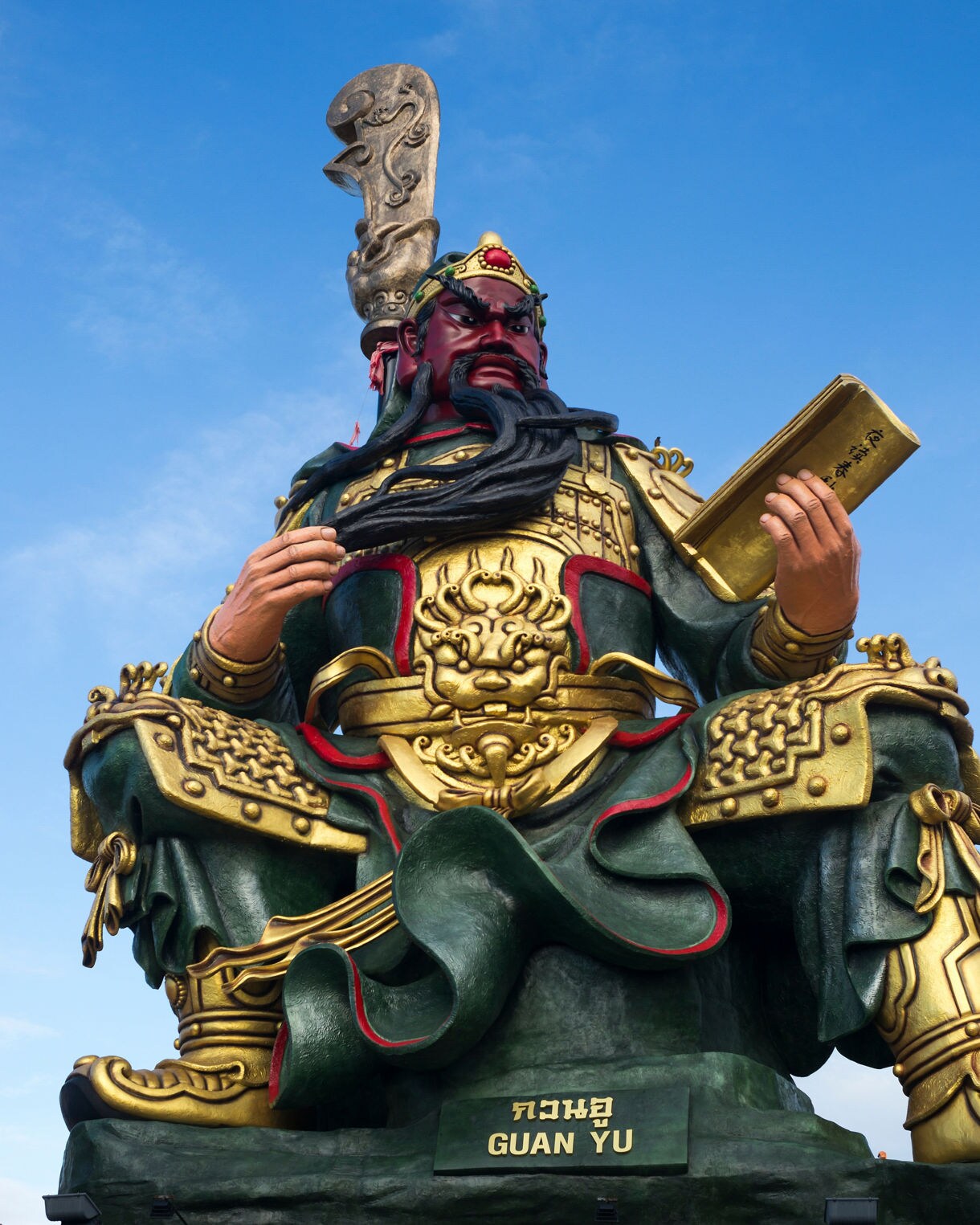 Massive statue of Guan Yu in ornate golden armor holding a book, set against a clear blue sky at Koh Samui’s Guan Yu Shrine.