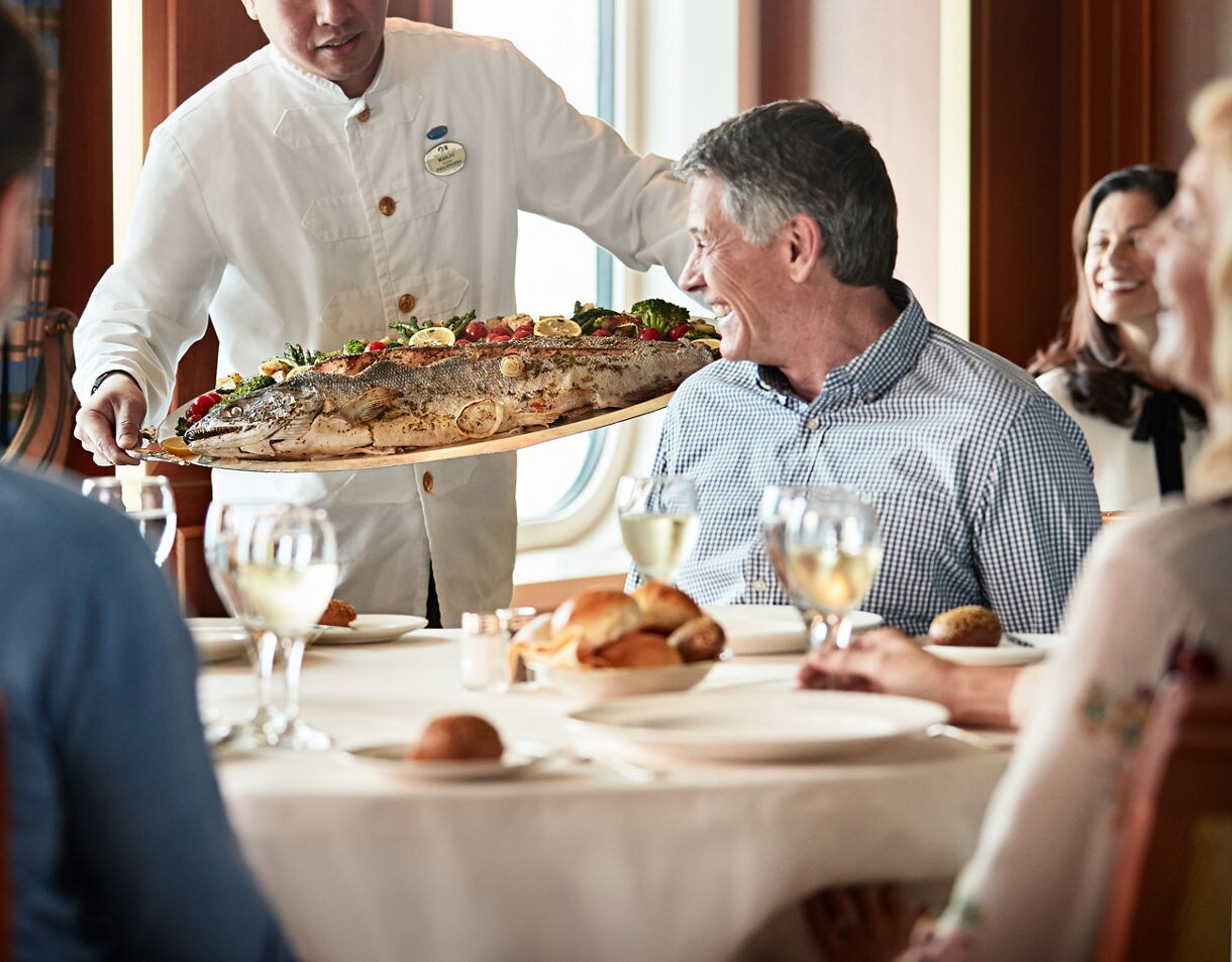 A waiter in a white uniform presents a large whole roasted fish garnished with vegetables to a group of seated adults. The setting is an upscale dining room with natural light and a round table set with wine glasses, bread rolls, and plates. The atmosphere is refined and social, with a focus on shared dining and hospitality. The color palette features warm wood tones and crisp whites, enhancing the elegant mood.